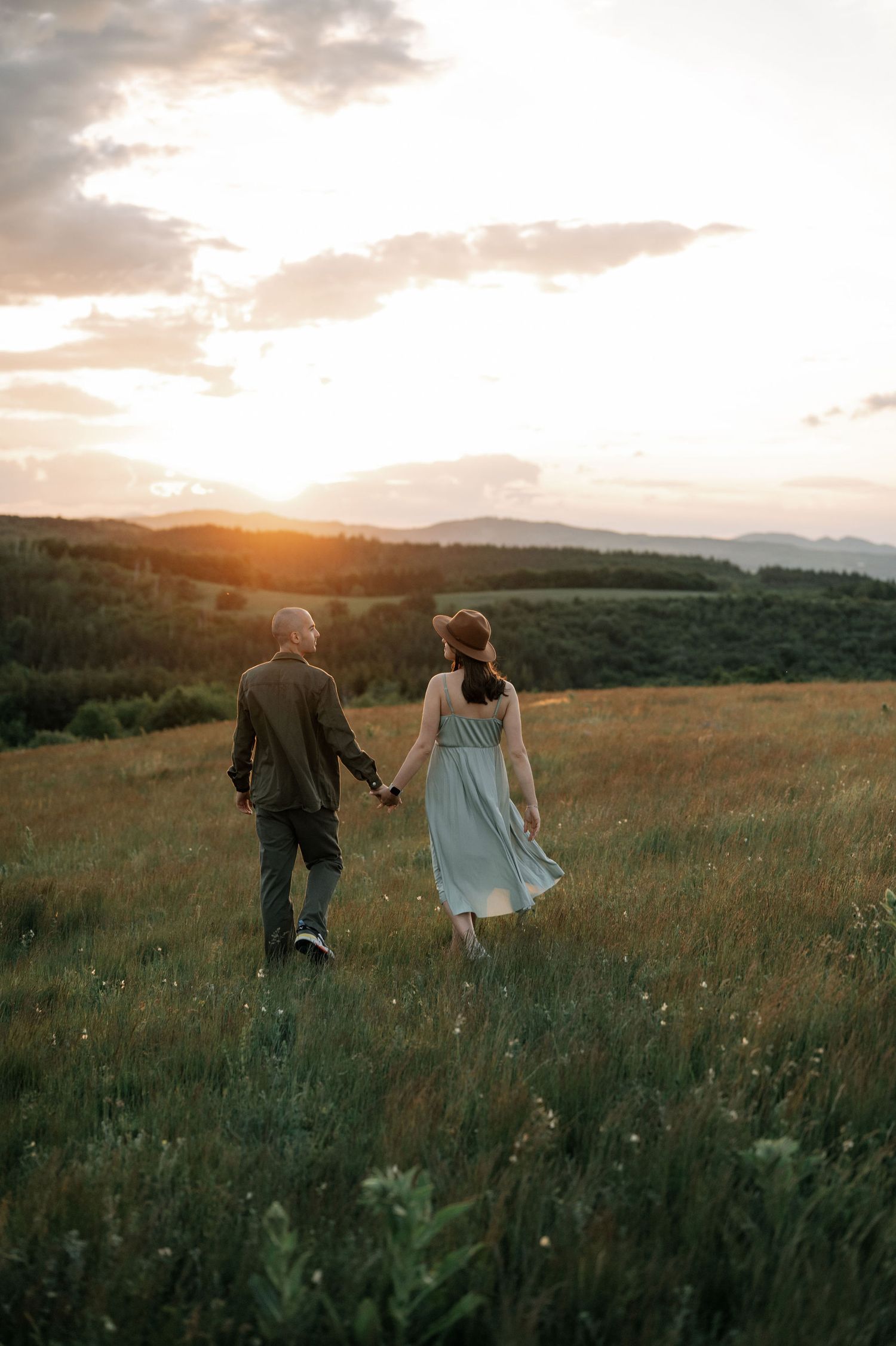 Couple holding hands walking through a green meadow at golden sunset with rolling hills in the background.