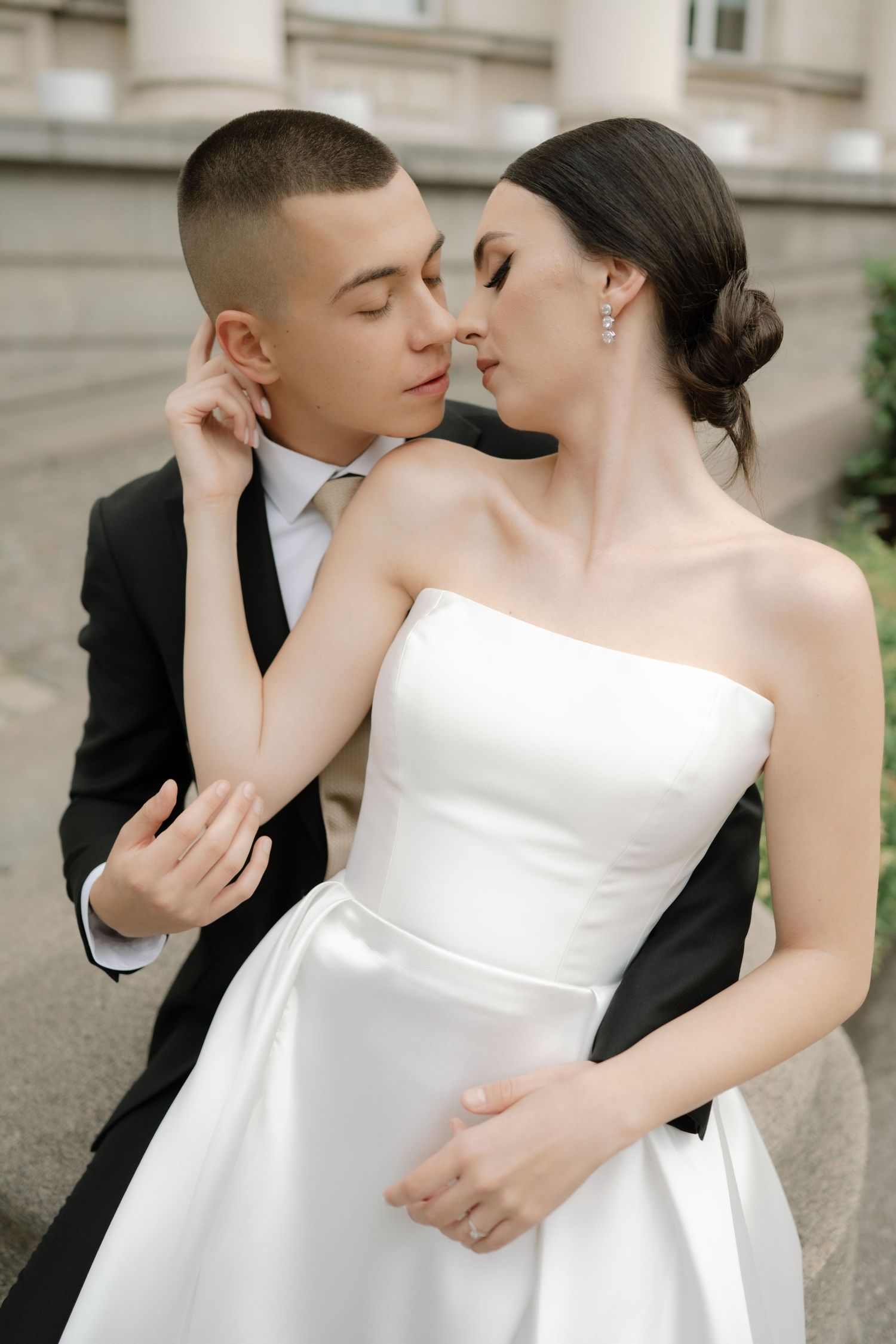 A romantic wedding couple in elegant attire share an intimate moment outdoors against a classical architectural backdrop.