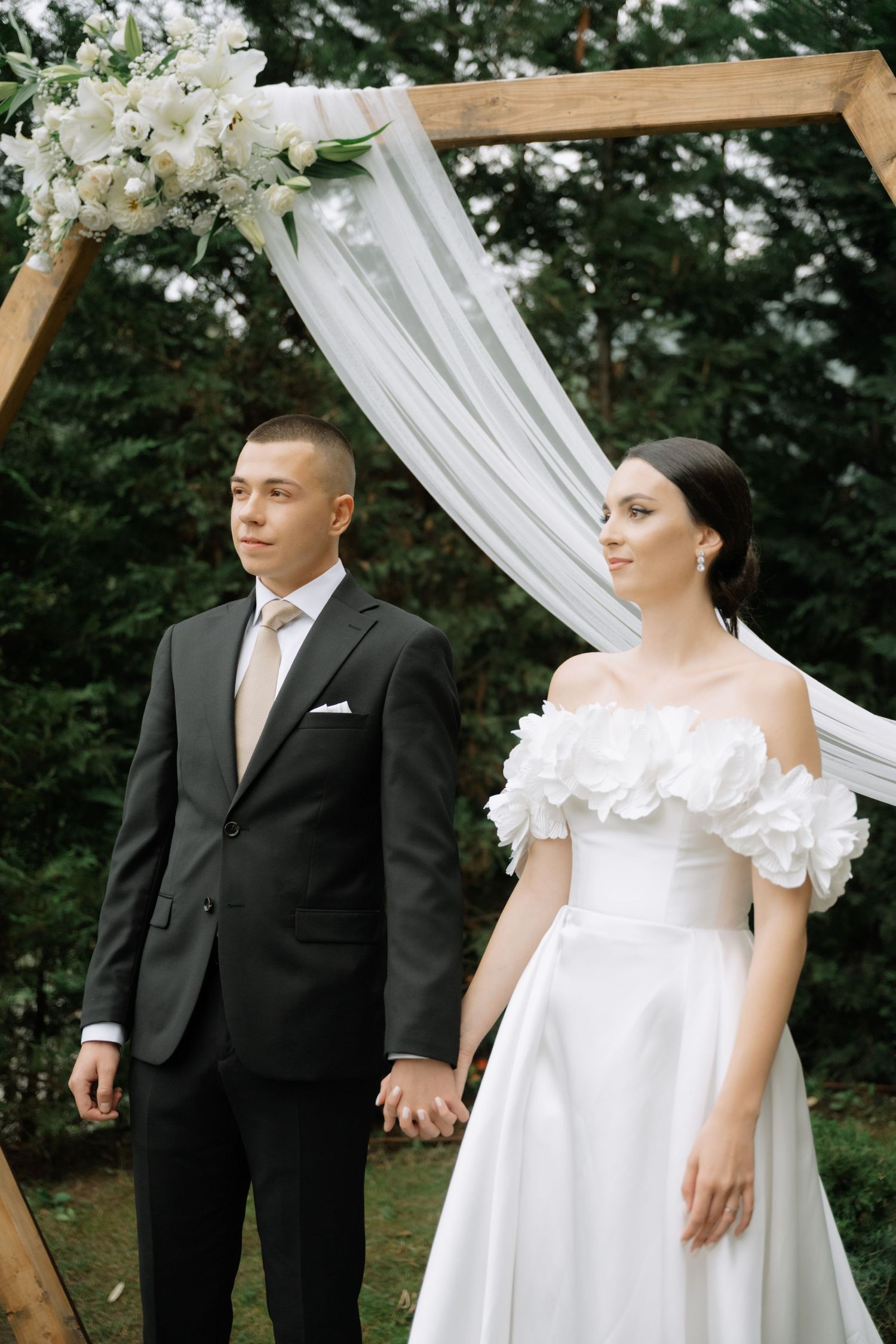 A couple in wedding attire pose together under a white draped arch during an outdoor ceremony.