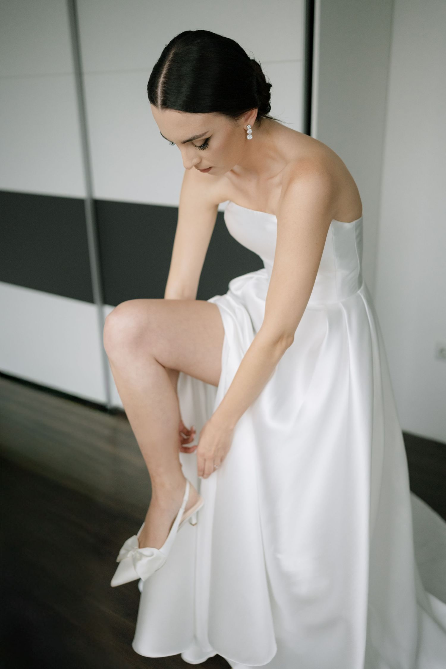 Elegant bride in strapless white gown adjusts her shoe while getting ready in minimalist room.