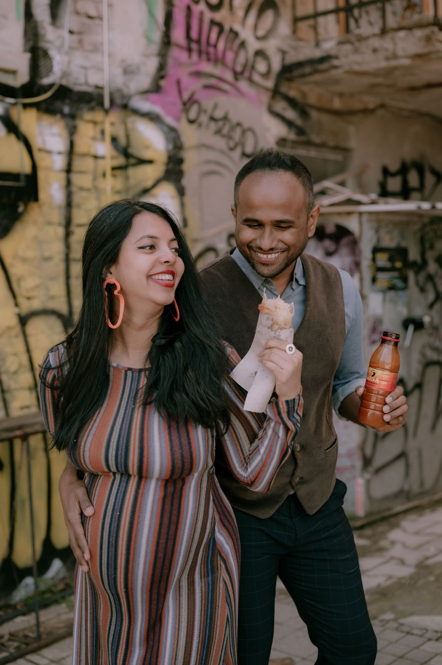 Happy couple laughing together in urban alley with colorful graffiti wall, woman in striped dress, man holding juice bottle.