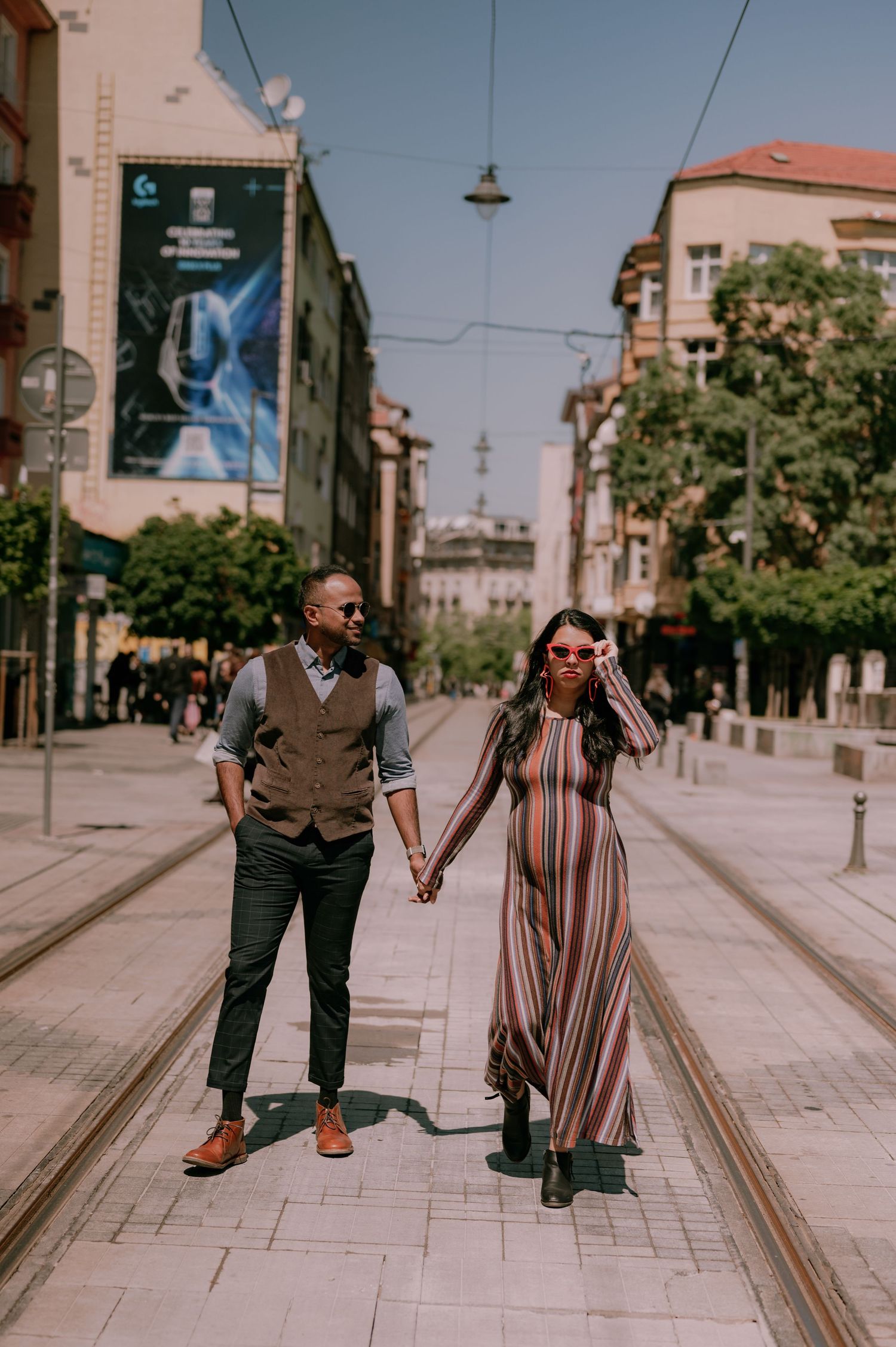 Couple holding hands walking on city street, man in vest and woman in striped maxi dress with sunglasses.