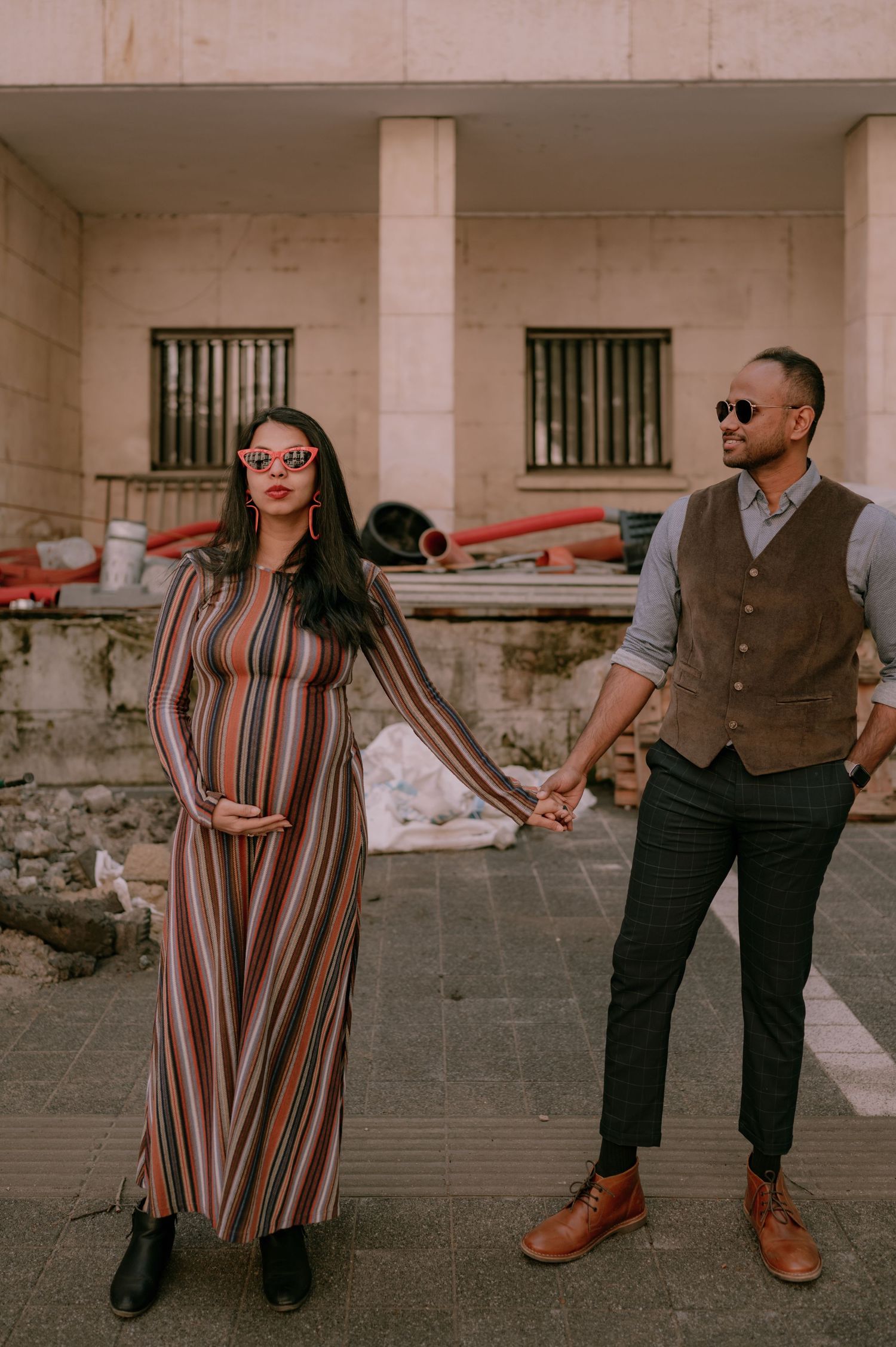Pregnant woman in striped maxi dress holds hands with man in vest and slacks outdoors near a building.
