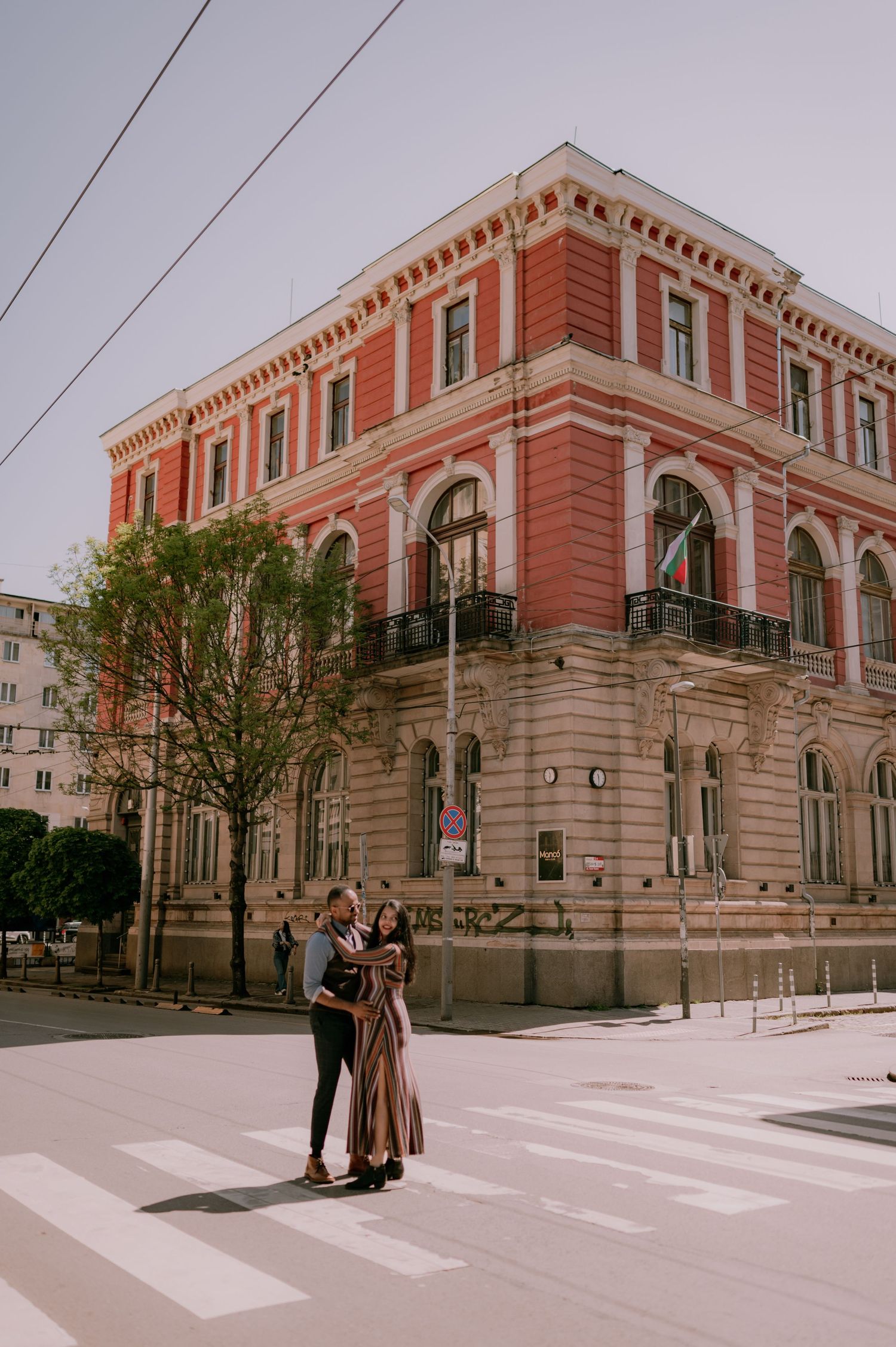 Couple embracing on a crosswalk in front of a grand red historic building on a sunny day.