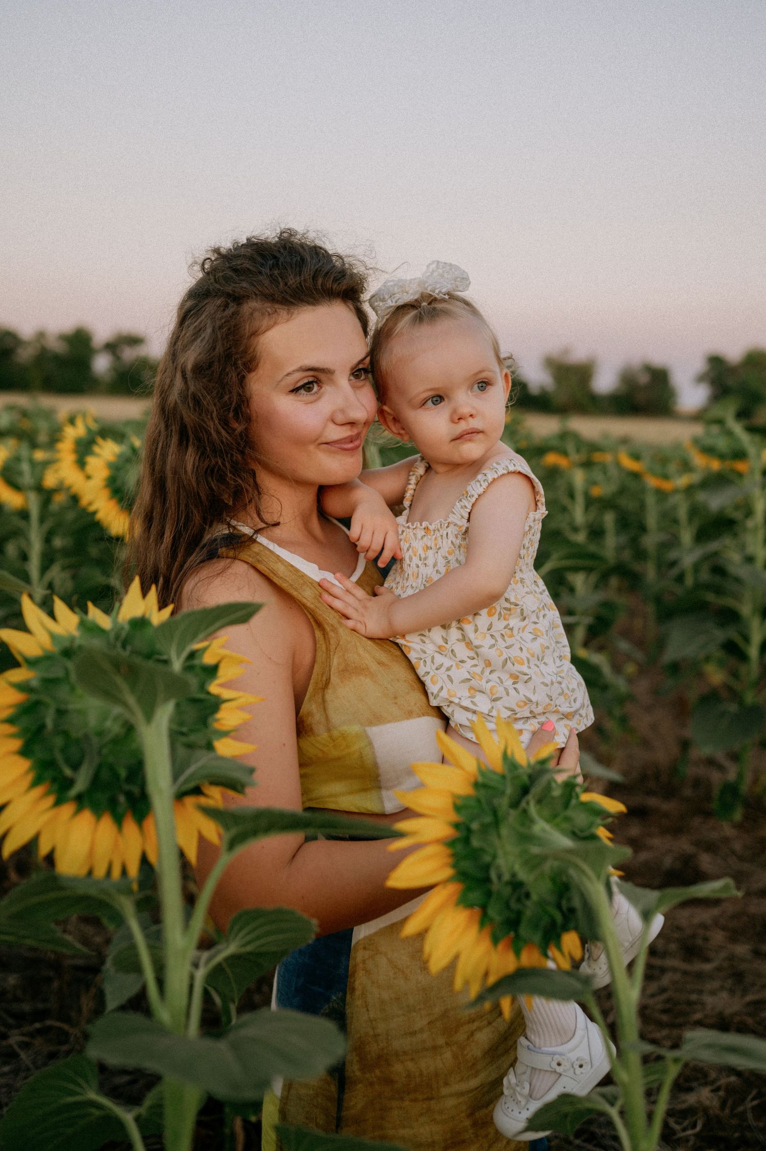 Mother holding baby daughter in sunflower field at sunset, both wearing yellow outfits.