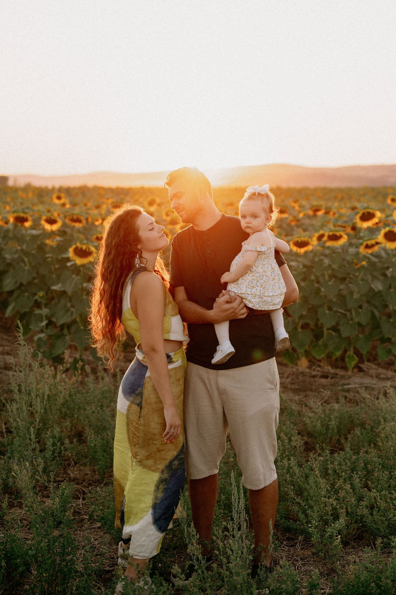 A family of three poses in a sunflower field at golden hour, with parents smiling at their baby girl.