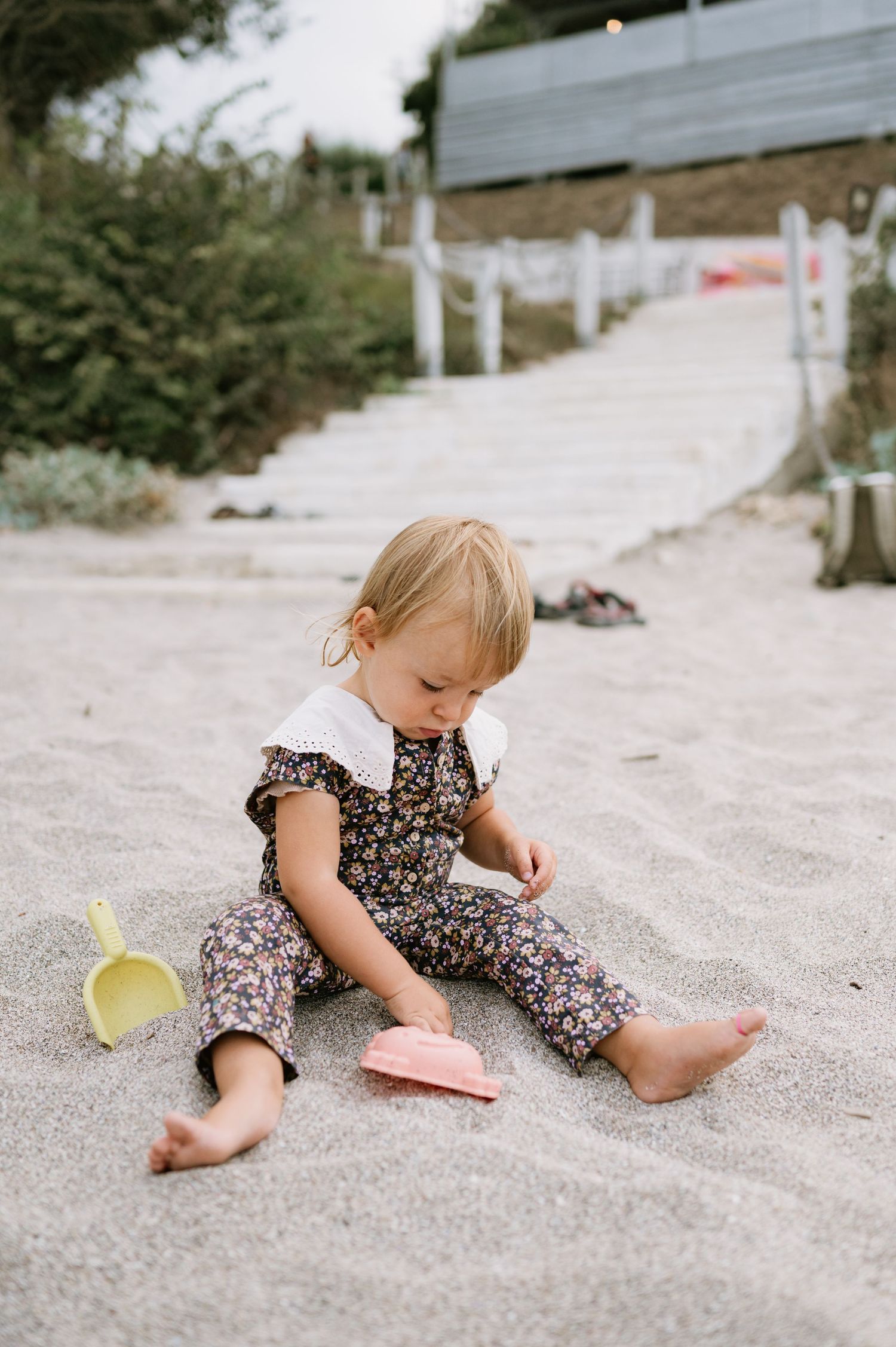 Toddler girl in floral romper sitting barefoot on sandy beach playing with pink and yellow toy shovel and bucket.