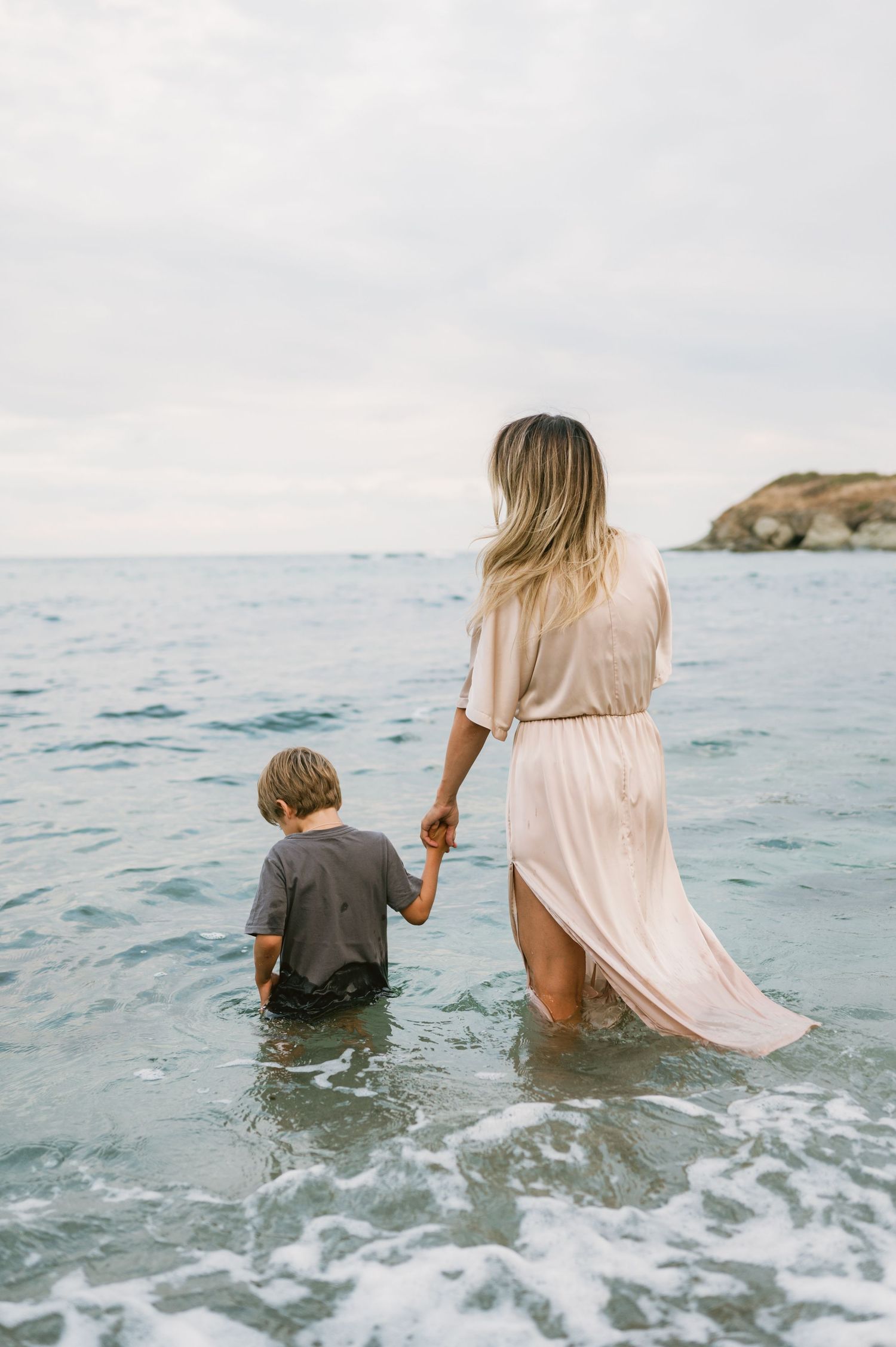 A mother and young child wade hand-in-hand into the ocean waves, viewed from behind on a cloudy day.
