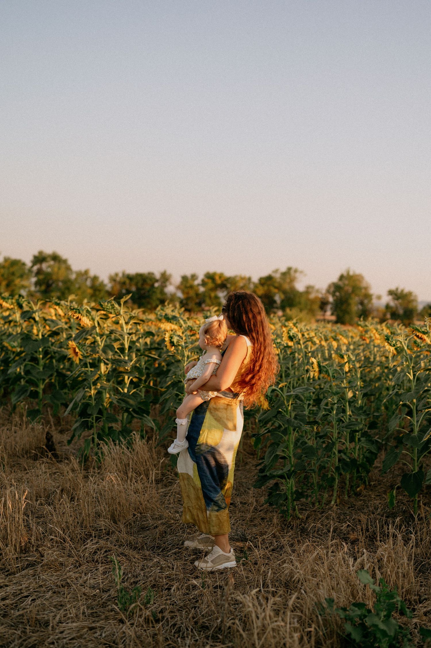 A mother holds her toddler in a sunflower field at golden hour, wearing tie-dye pants, surrounded by lush green blooms.