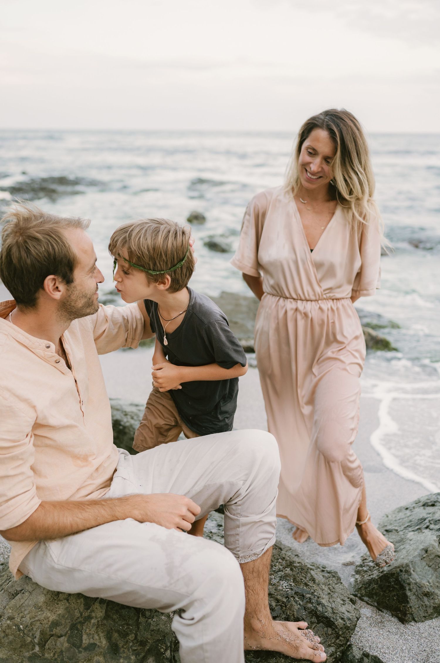 Family of three enjoying a relaxed beach moment at dusk, with parents and child sitting on rocks by the ocean.