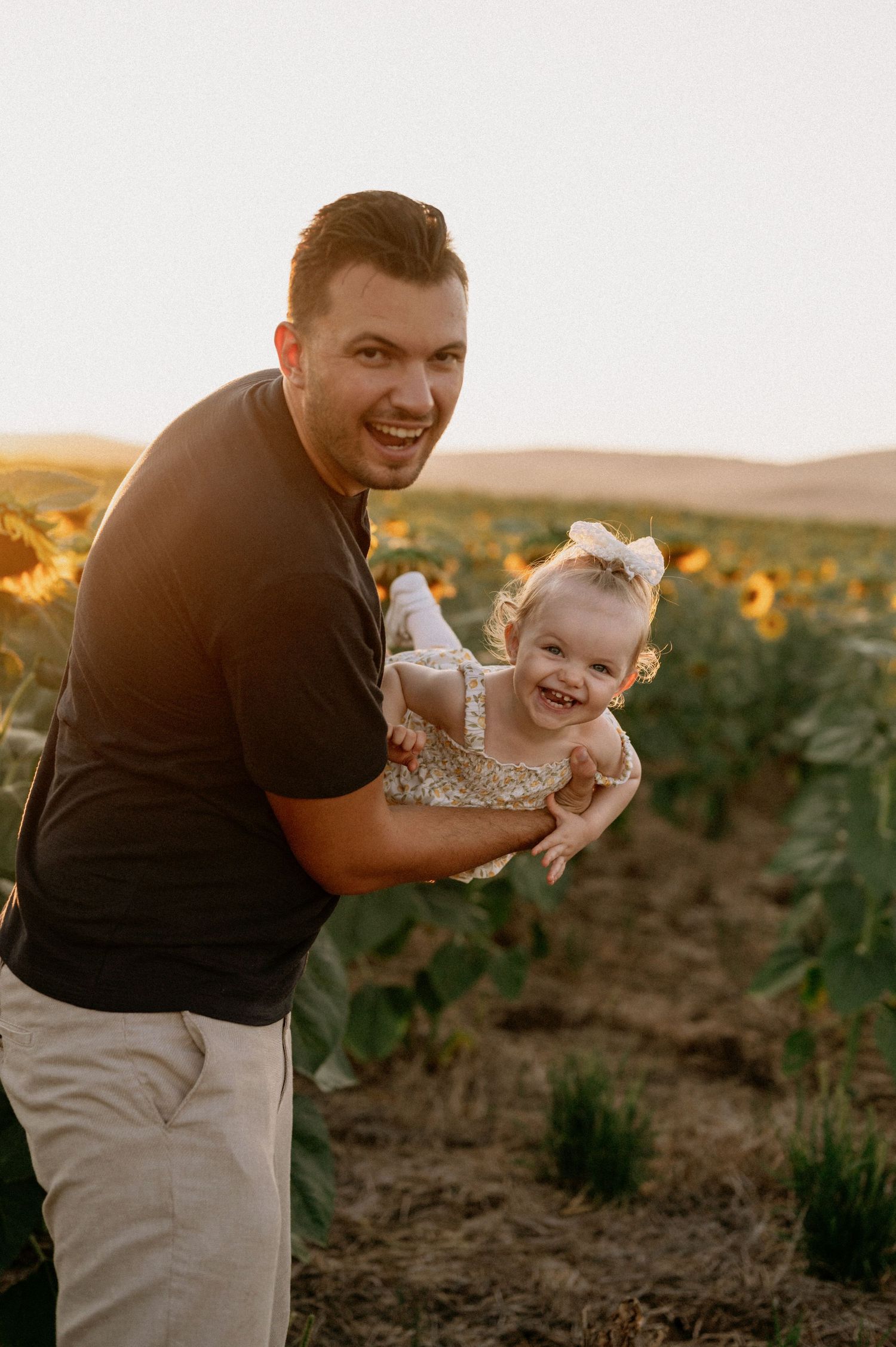 A joyful father holds his laughing toddler daughter in a sunflower field during golden hour sunset.