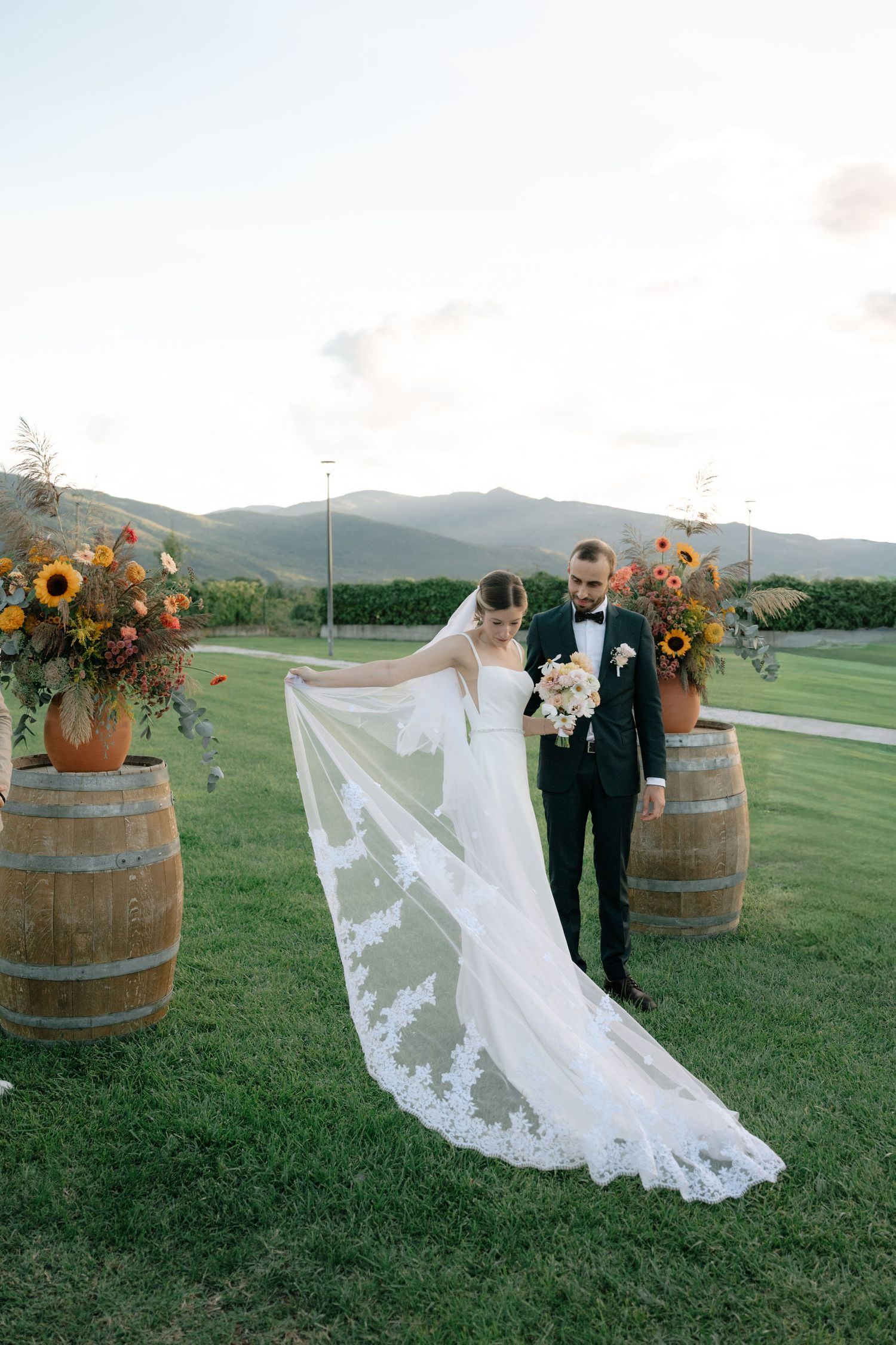 Newlywed couple poses with rustic wine barrels and sunflower decorations in a scenic mountain landscape at sunset.