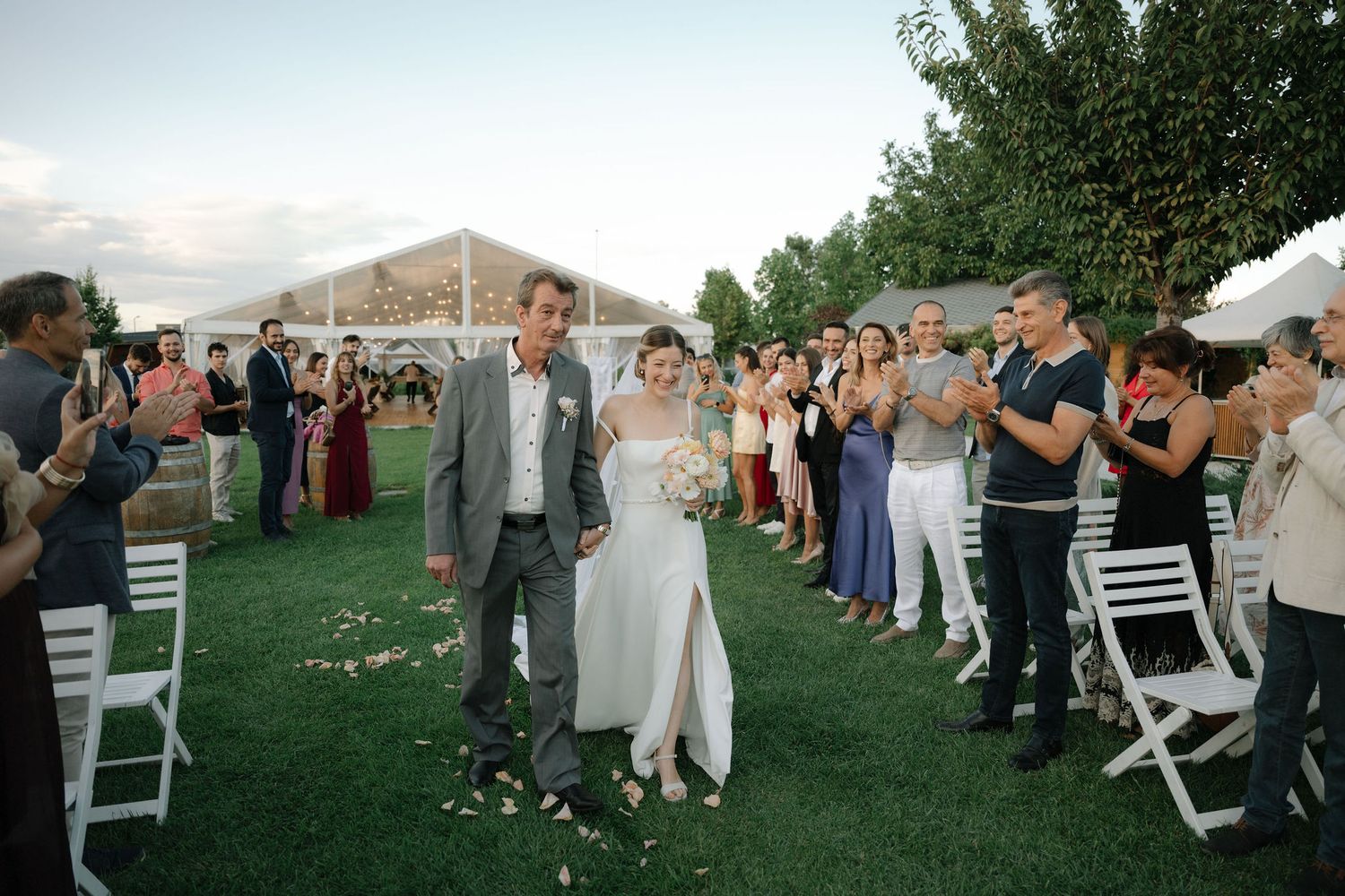 Wedding guests dance together on a grassy lawn during an outdoor reception celebration at sunset.