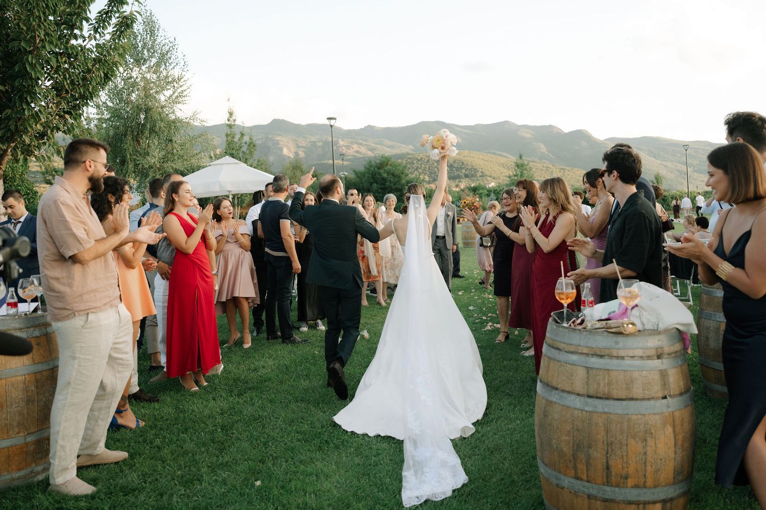 Wedding guests dance in a circle during an outdoor reception celebration with wine barrels and rolling hills in background.