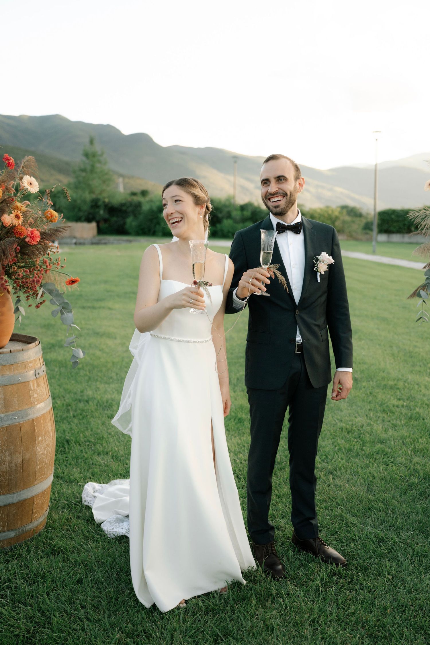 Newlywed couple toasts with champagne glasses against scenic mountain backdrop on grassy lawn at outdoor wedding.