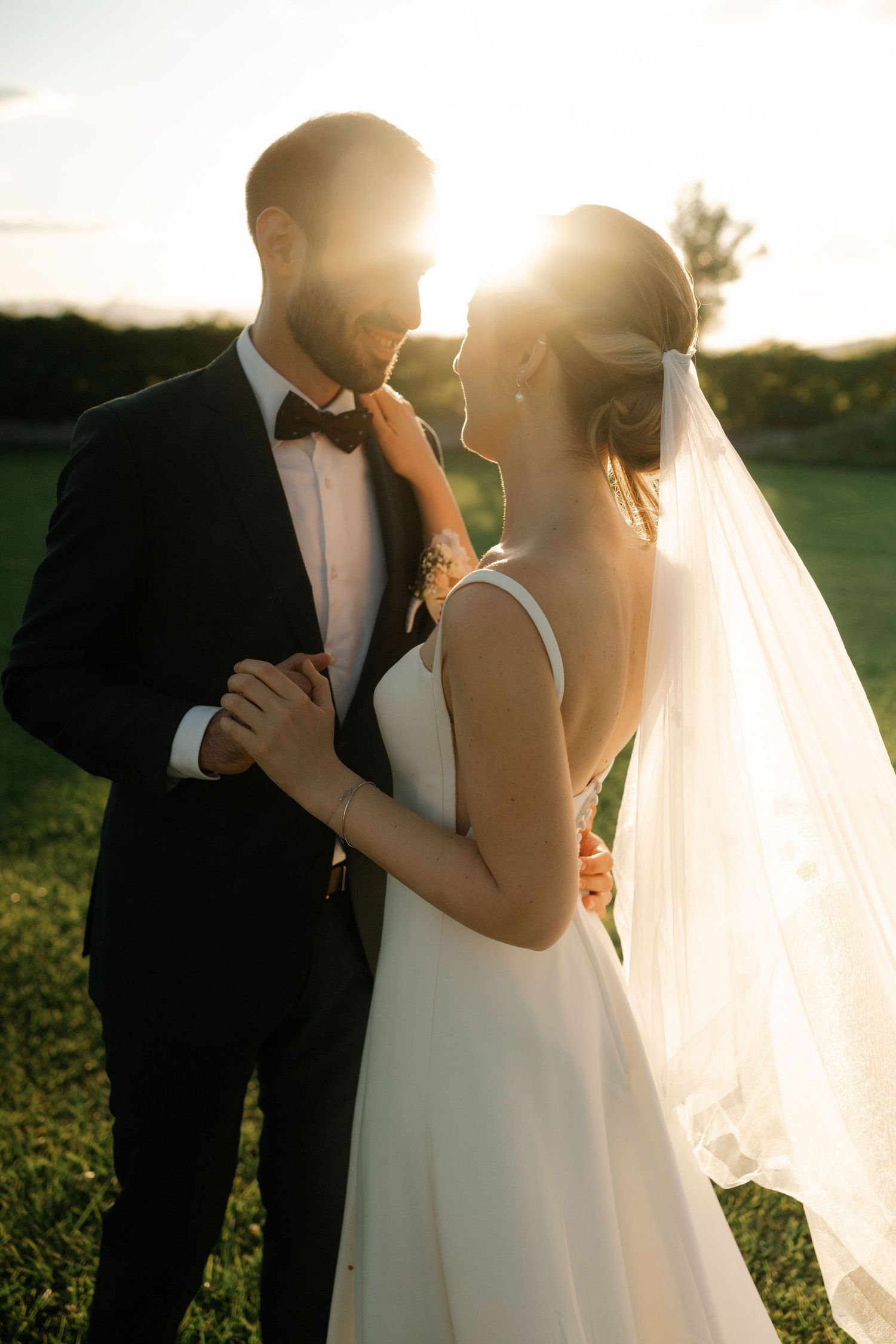Romantic wedding couple embraces in golden sunlight as bride's veil flows in gentle breeze during sunset photoshoot.