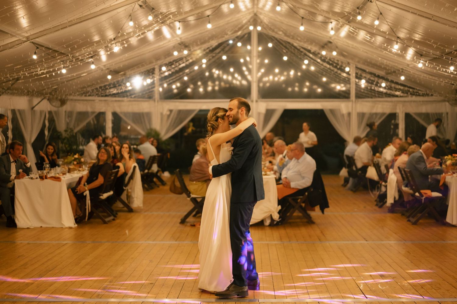 A couple shares their first dance under twinkling lights in a romantic wedding tent reception with guests watching.