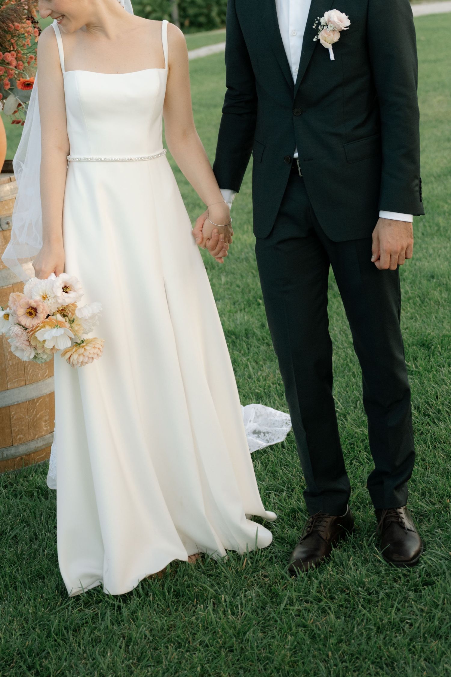 Close-up details of a bride in white dress and groom in black suit standing together on green grass during wedding ceremony.