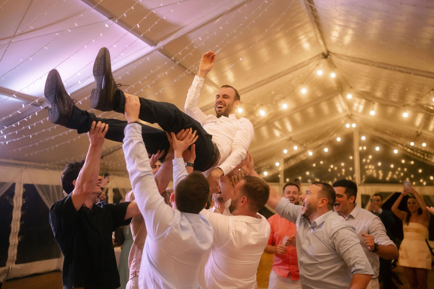 Guests celebrate at a wedding reception by lifting someone up during a dance party under a tent with string lights.