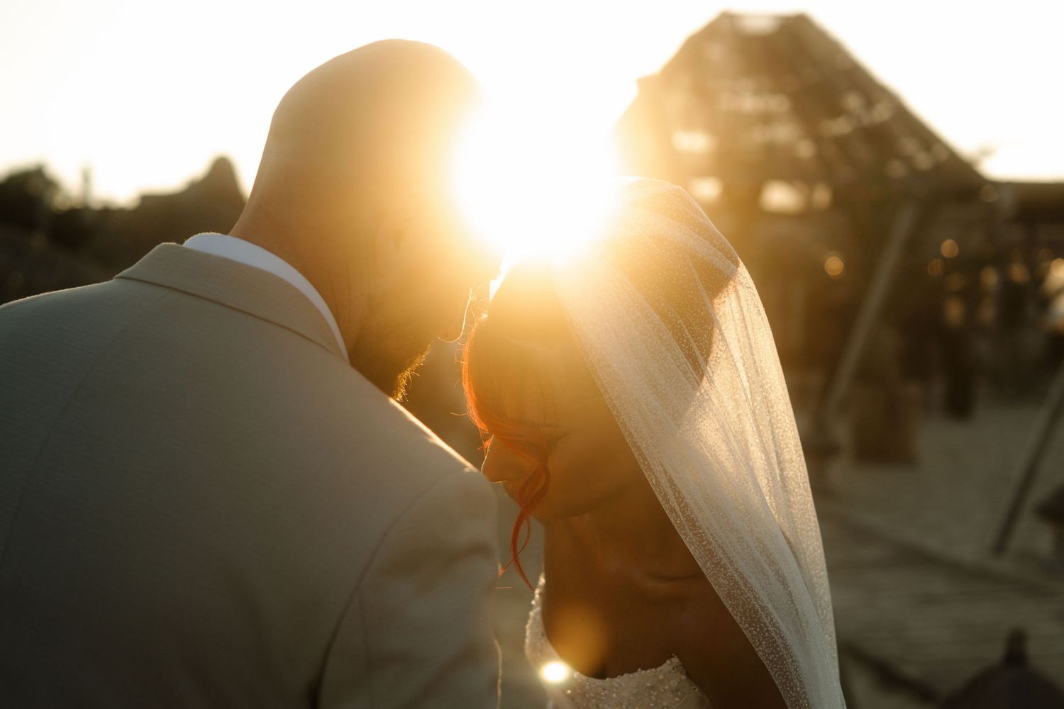 A bride and groom share an intimate moment at sunset, with sunlight creating a warm backlit glow through her veil.