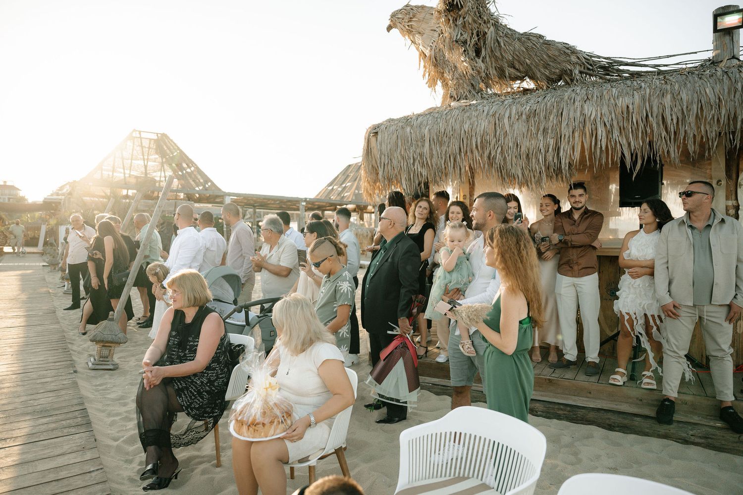 Guests in white and pastel attire gather under a thatched-roof beach pavilion during a sunset celebration.