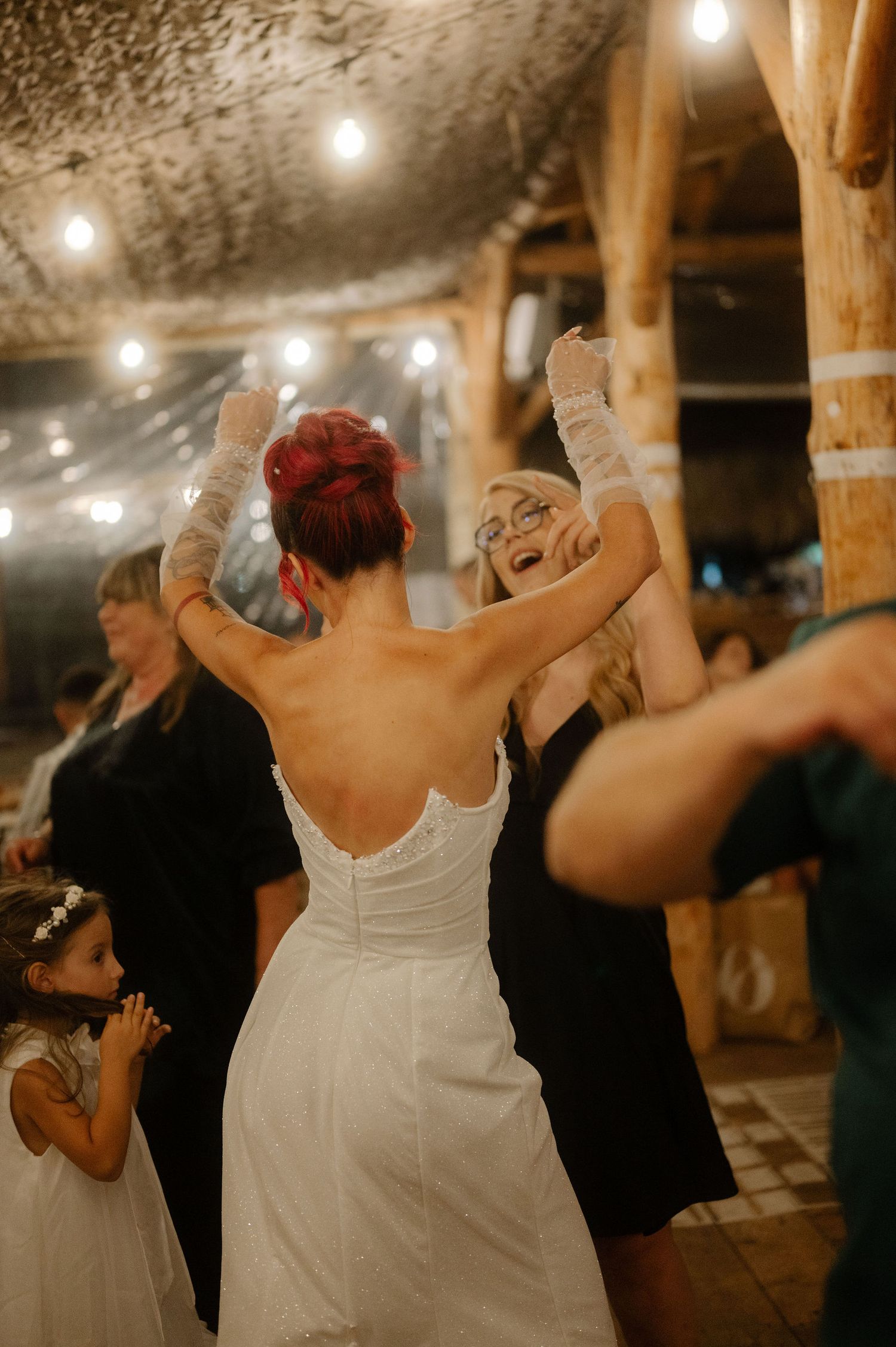 Wedding guests dancing and celebrating at a rustic barn venue with string lights overhead.