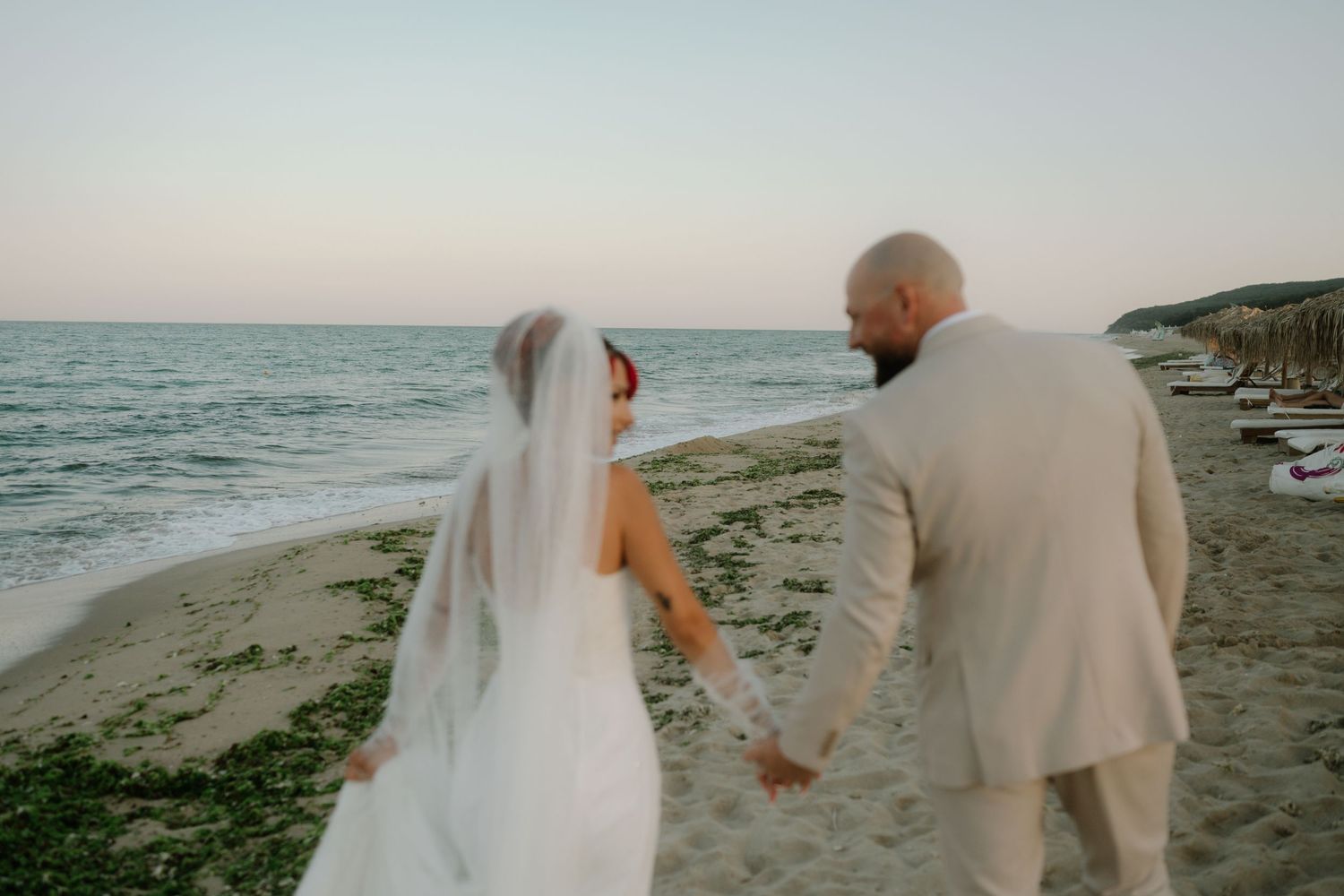 A couple in wedding attire walks along a scenic beach shoreline at sunset.