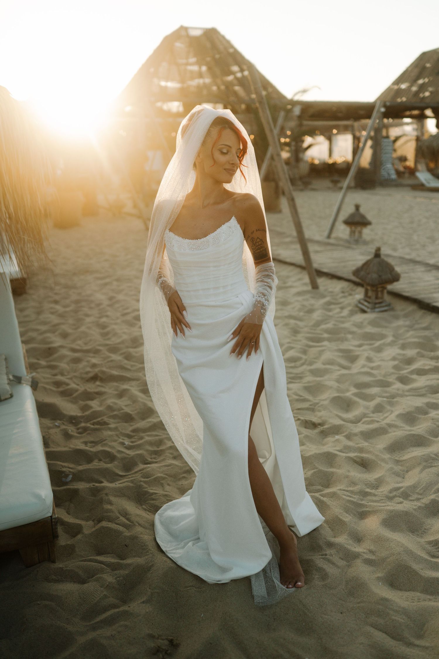 Bride in a sleek white wedding dress poses gracefully on a sandy beach at sunset.