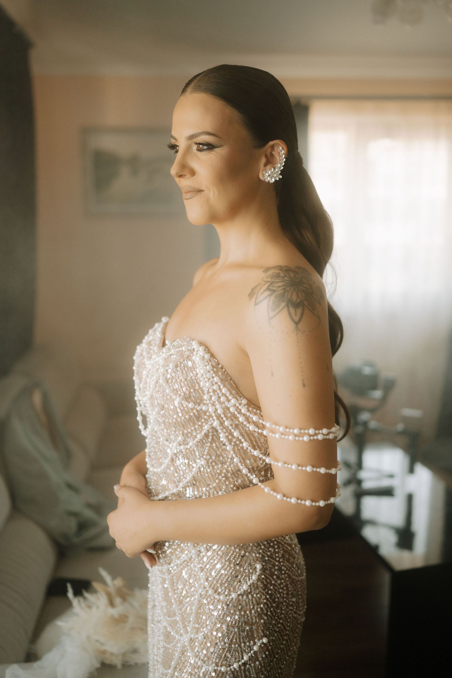 A bride in a sparkling beaded off-shoulder wedding gown poses during preparations in a softly lit room.