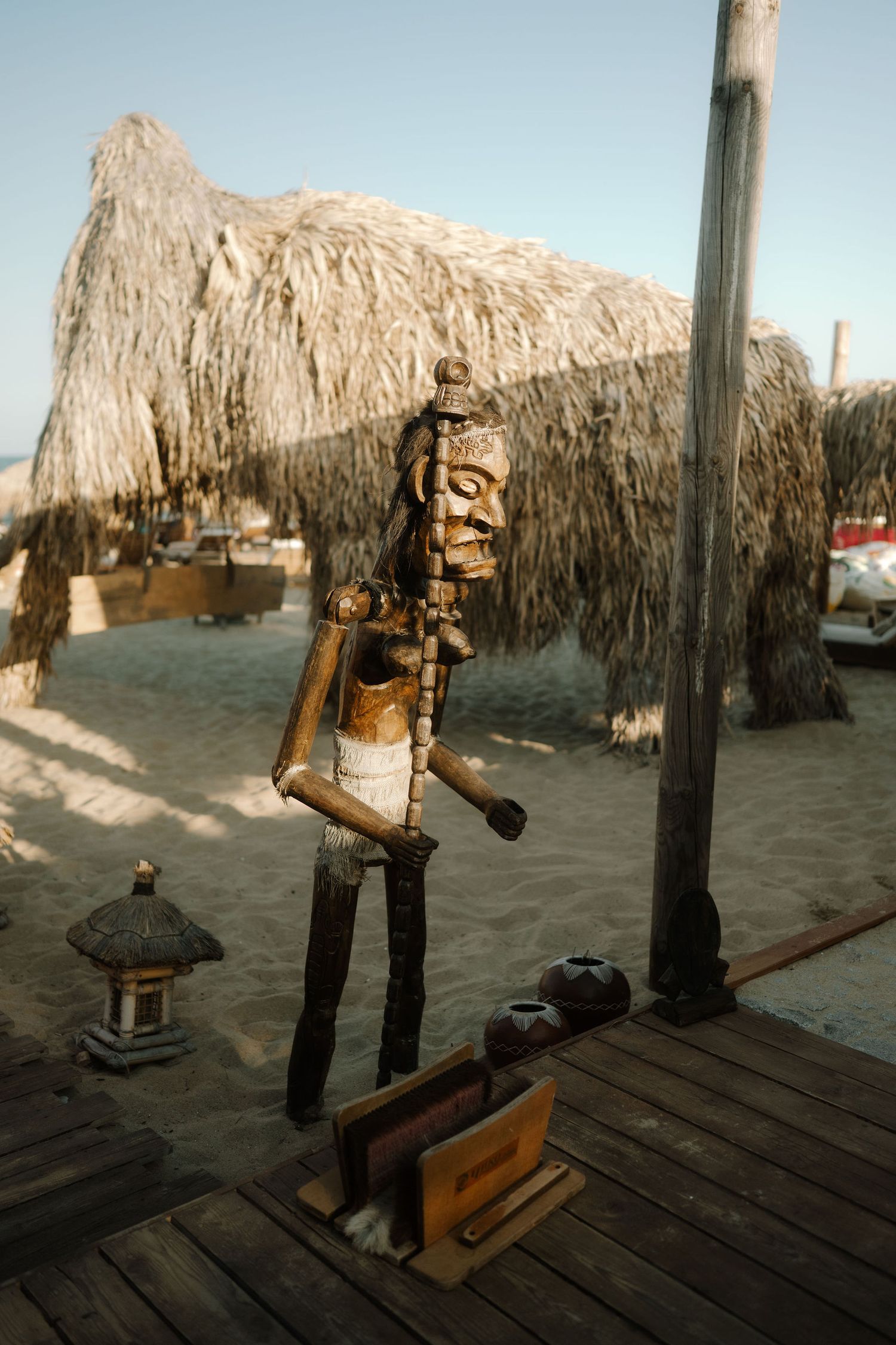 A wooden carved tiki statue stands on a deck with a thatched-roof structure and small lantern in the background.
