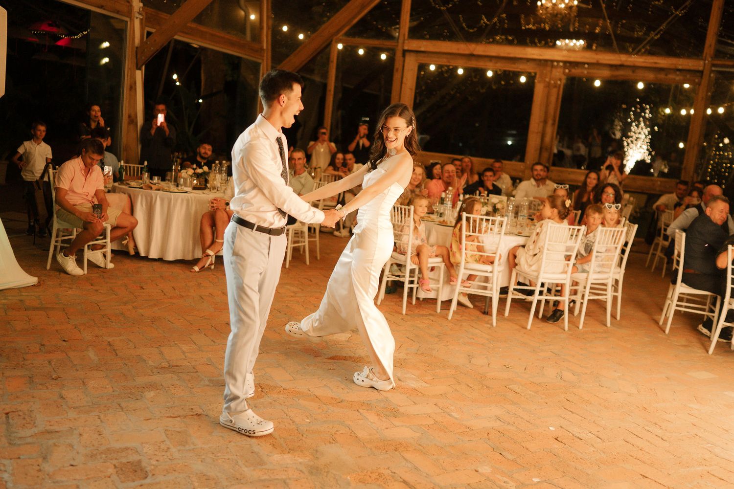 A couple shares their first dance at an elegant indoor reception with string lights and white chairs in the background.