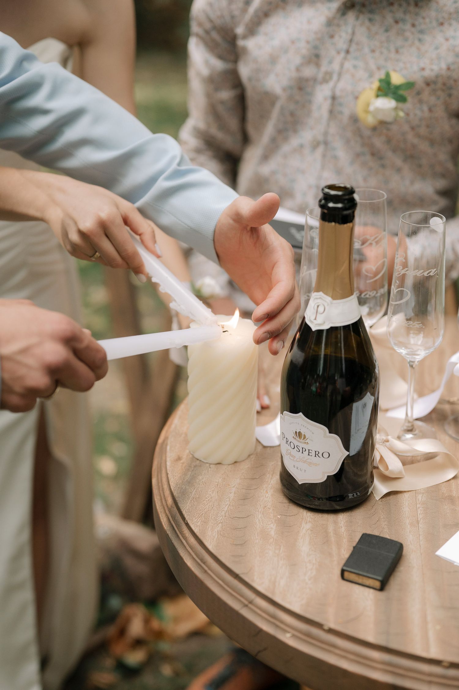 Hands lighting a white candle on a round table with champagne bottle and empty glasses during an outdoor celebration.