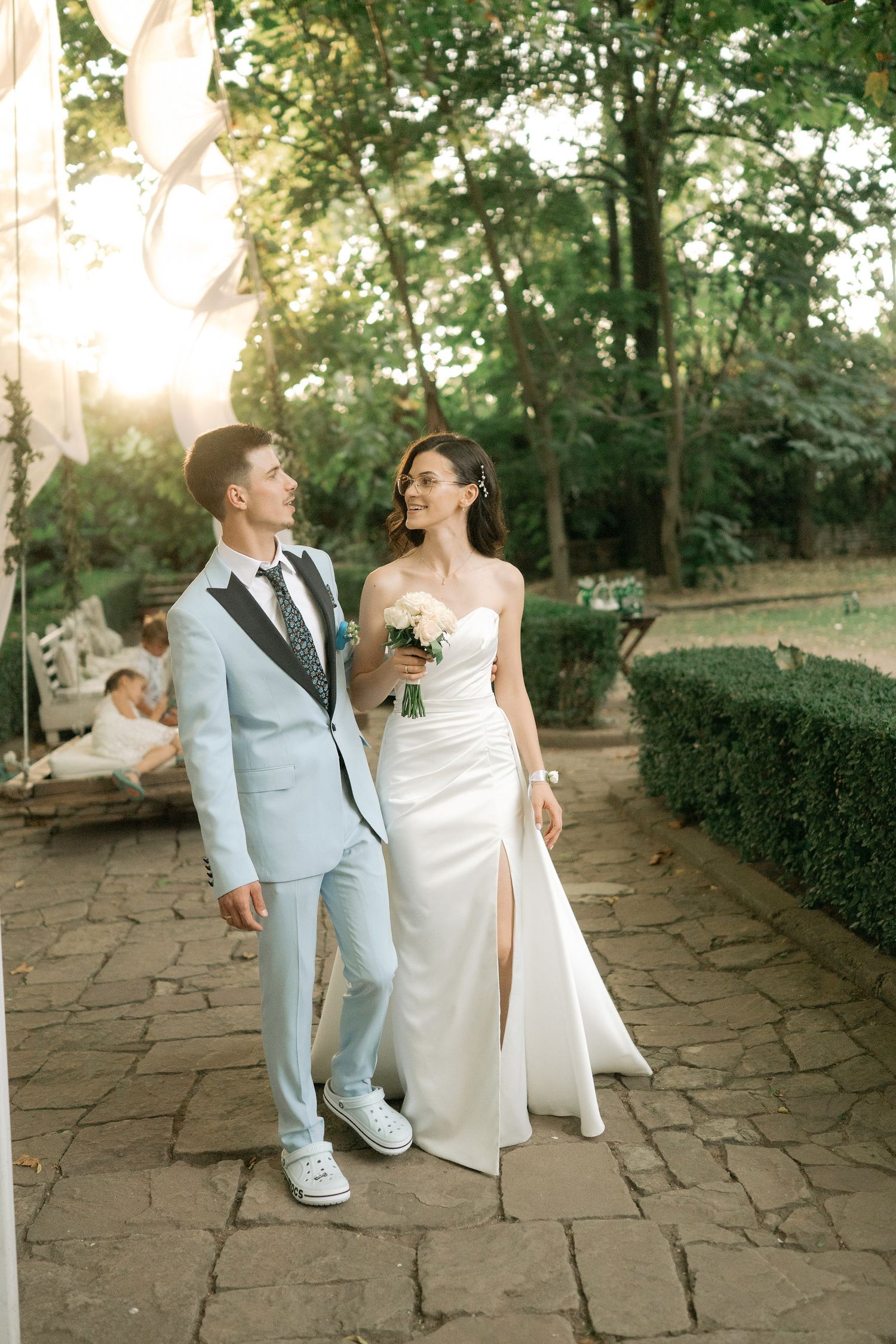 A couple in wedding attire walking and laughing together on a stone path surrounded by greenery at sunset.