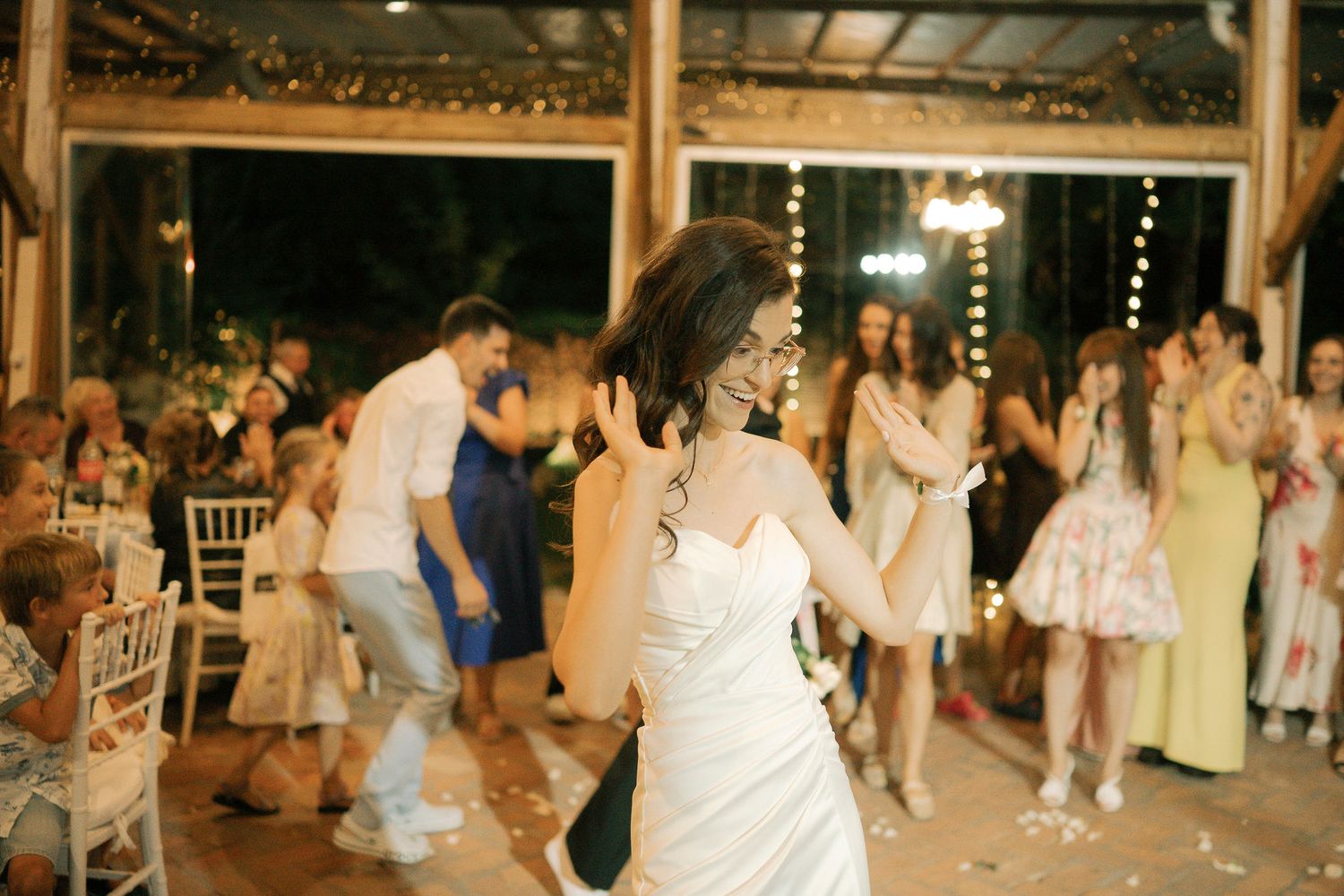 Wedding guests dance and celebrate on a wooden dance floor under twinkling lights at an evening reception.