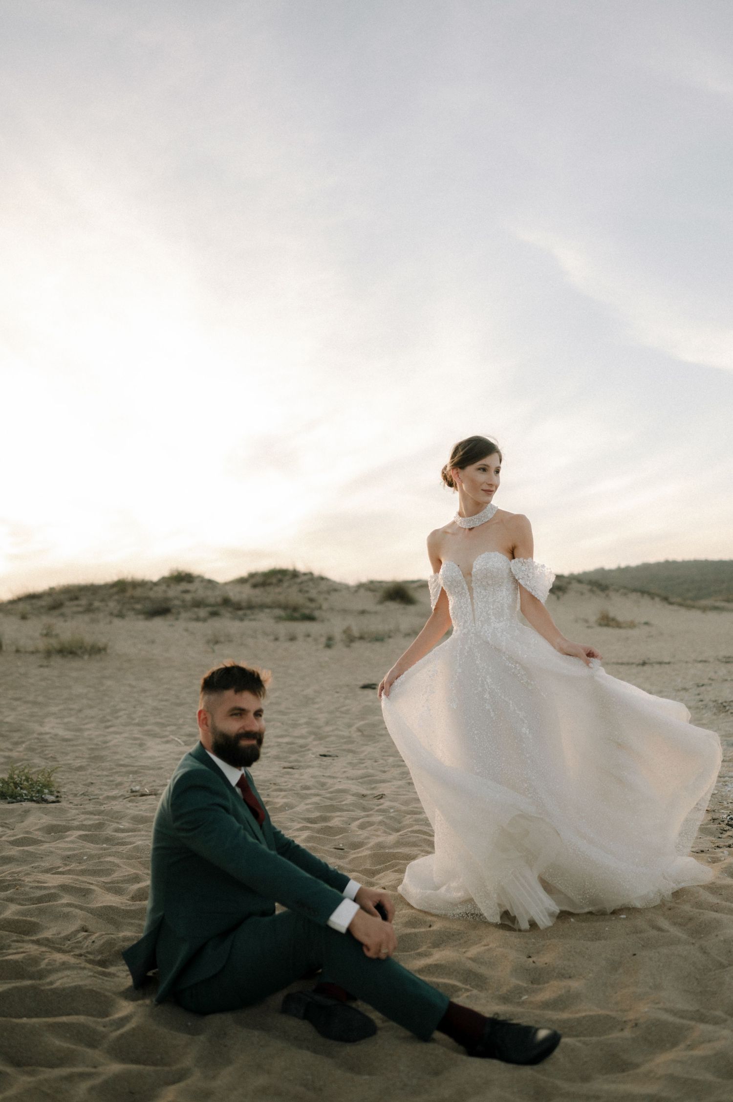 Groom sits on sand dunes while bride in flowing lace off-shoulder wedding gown stands behind him at sunset.