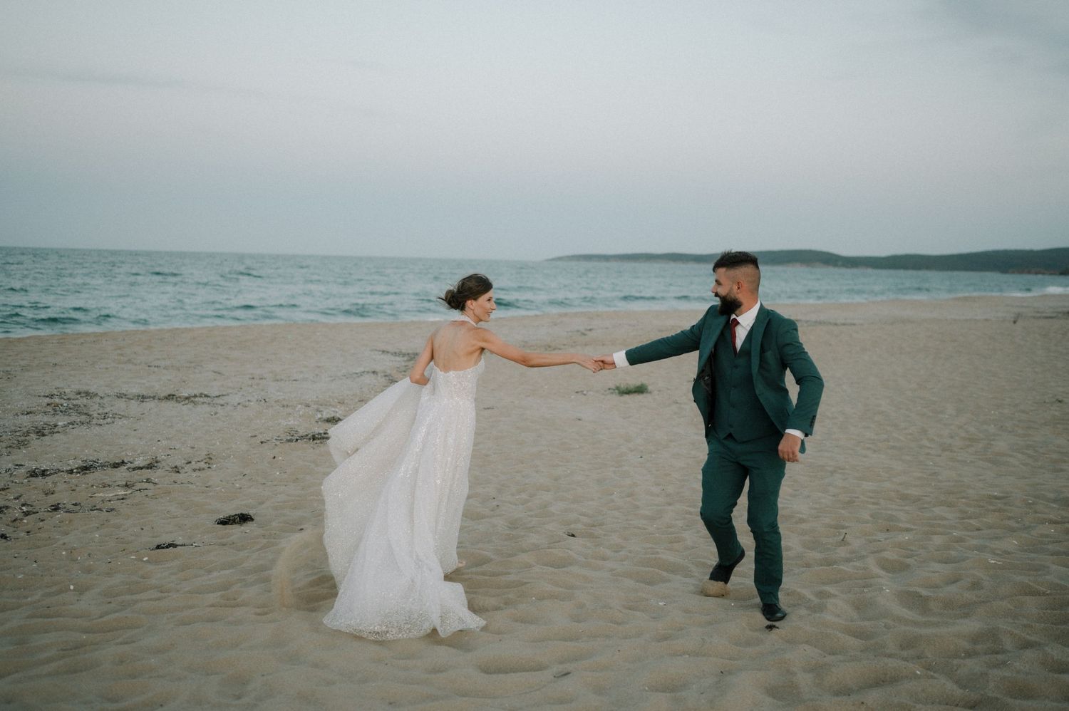 Bride and groom hold hands walking on a sandy beach at dusk, bride in white gown, groom in teal suit.