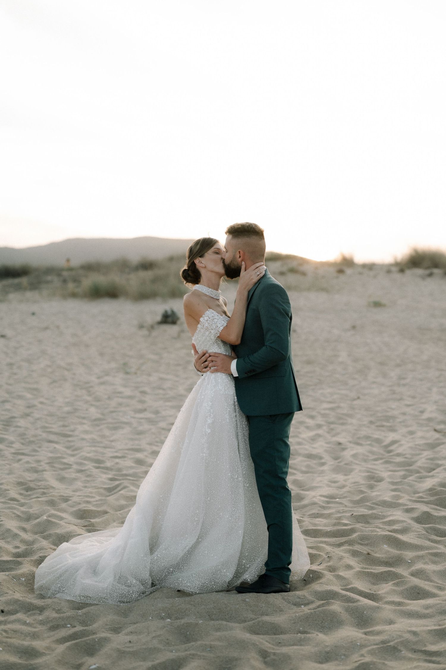 A bride and groom share a romantic kiss on a sandy beach at sunset, with the bride in a flowing white gown.