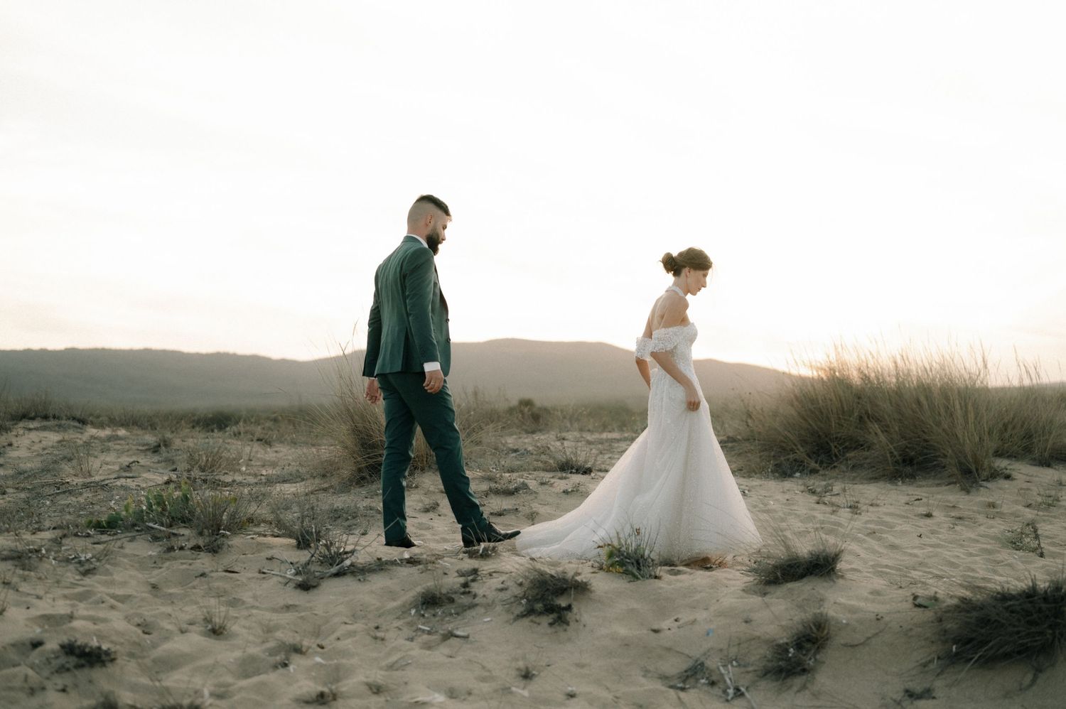 A bride in a white gown and groom in a dark suit walk through a sandy desert landscape at sunset.