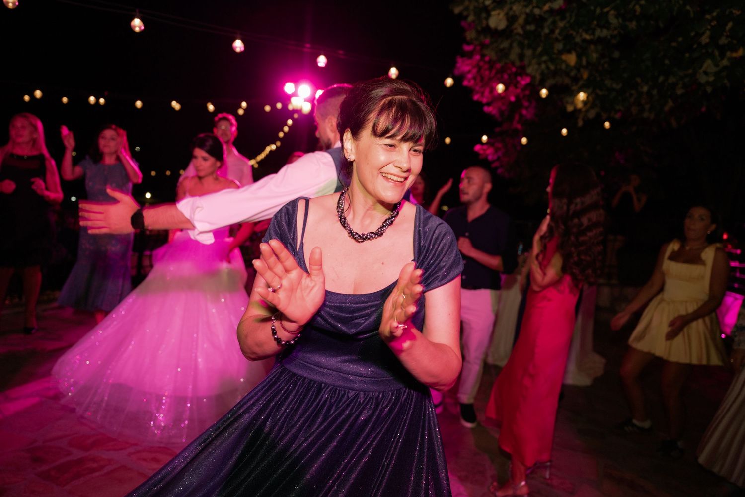 People dancing and celebrating at a wedding reception under vibrant pink and purple lighting.