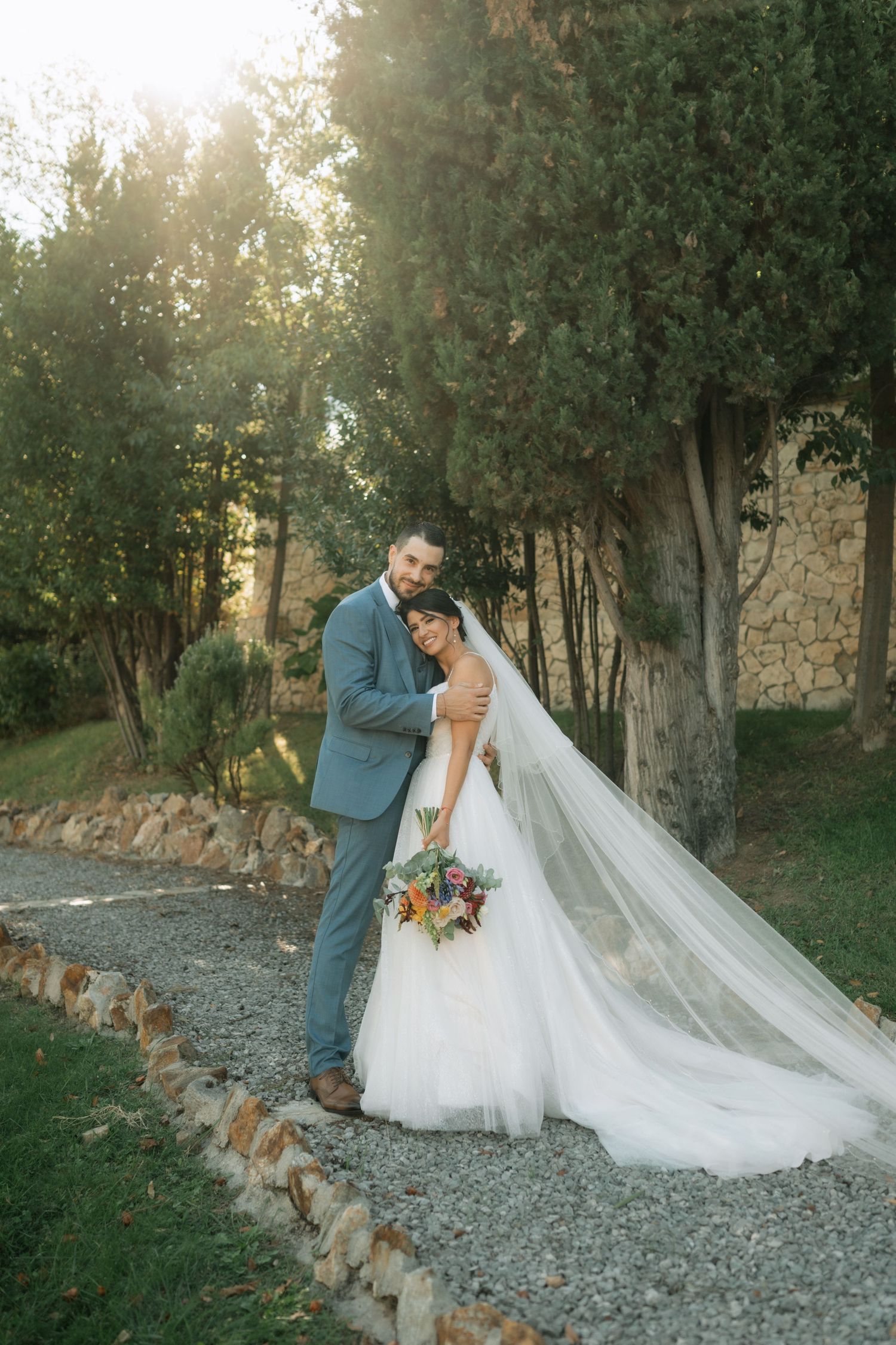 A couple in wedding attire share an intimate moment on a stone path surrounded by lush greenery at sunset.
