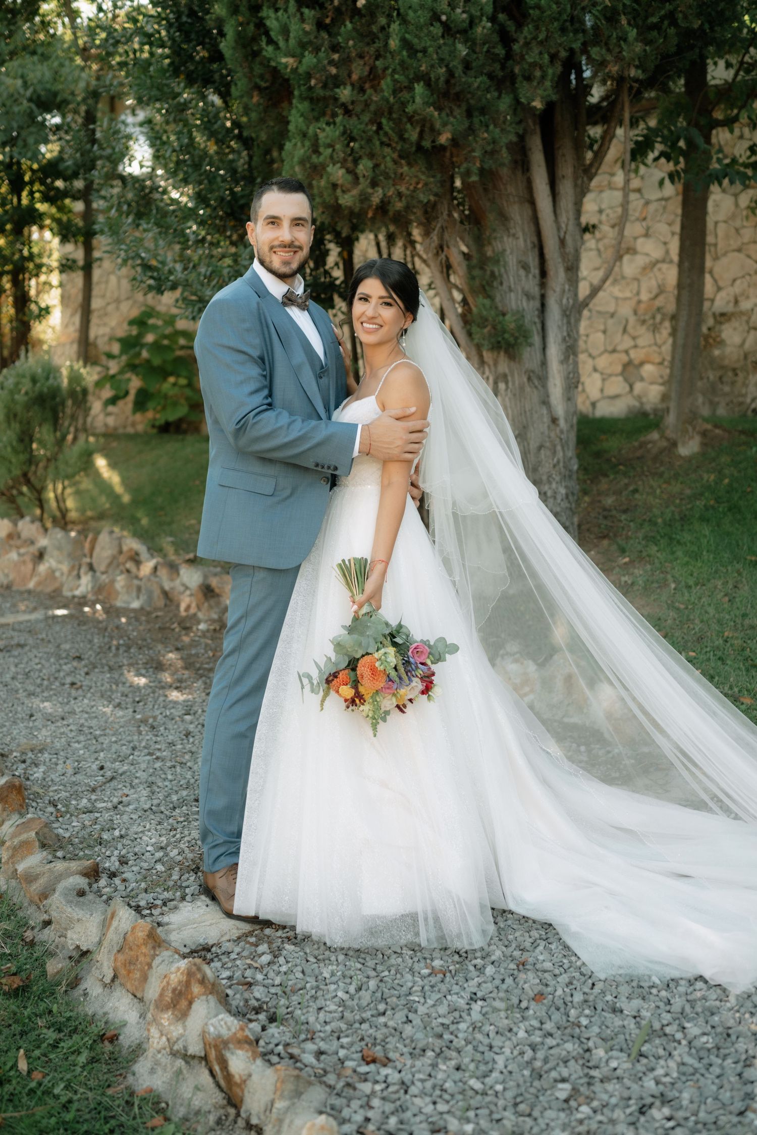 A couple poses outdoors in wedding attire with a long flowing veil and colorful bouquet on a gravel path near stone walls.