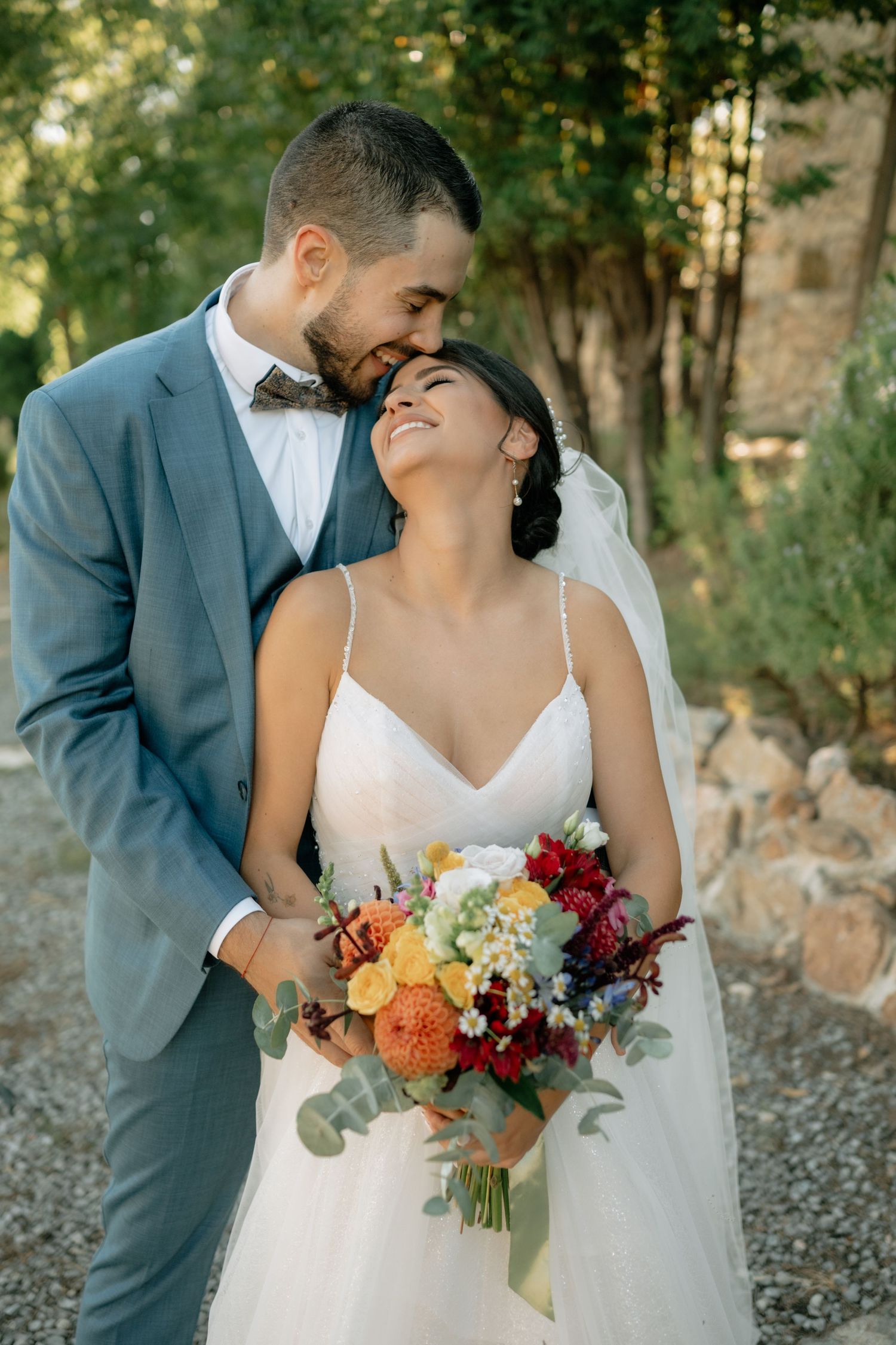A romantic wedding couple shares tender moments in a wooded outdoor setting with vibrant autumn bouquet details.
