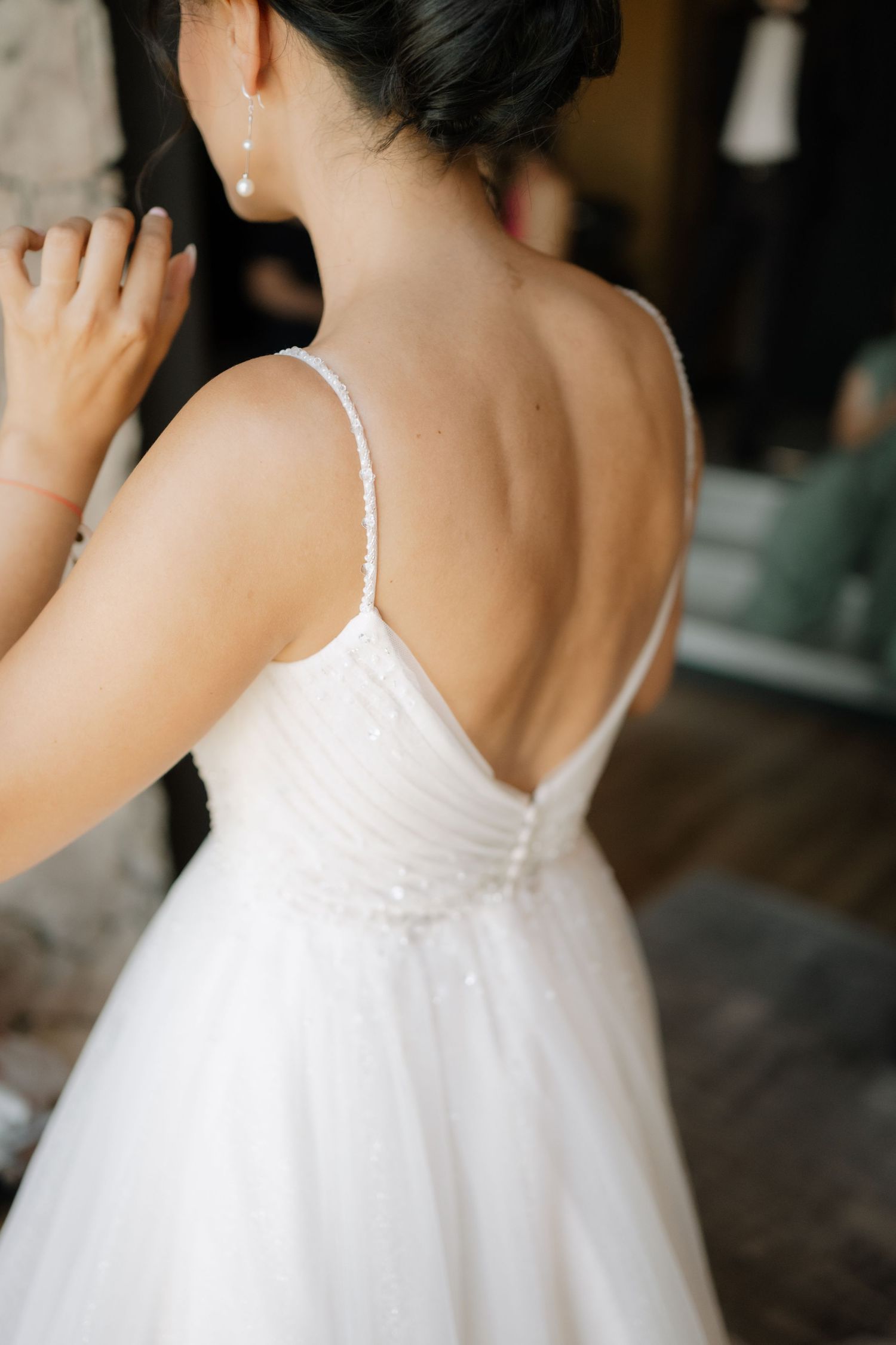 Back view of a bride in an elegant white wedding gown with thin straps and tulle skirt during wedding preparations.