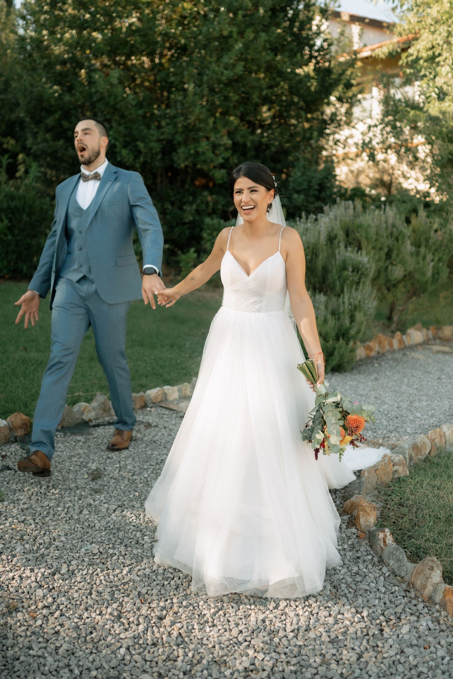A couple walks on a gravel path during their wedding, with the bride in white holding a bouquet and groom in blue suit.