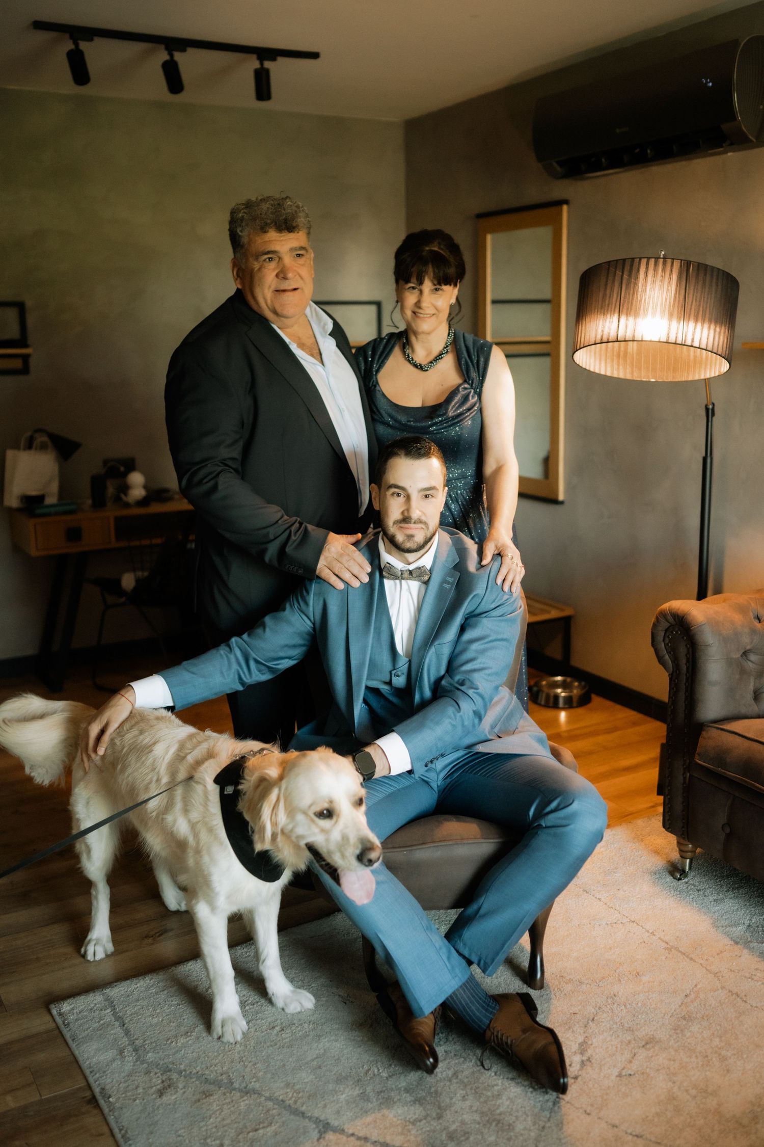 Two people in formal attire pose with their white dog on a beige carpet in a cozy living room setting.