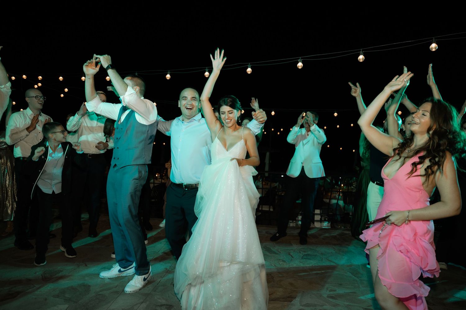 Wedding reception guests dance and celebrate on a dimly lit dance floor illuminated by colorful lights and string lights.