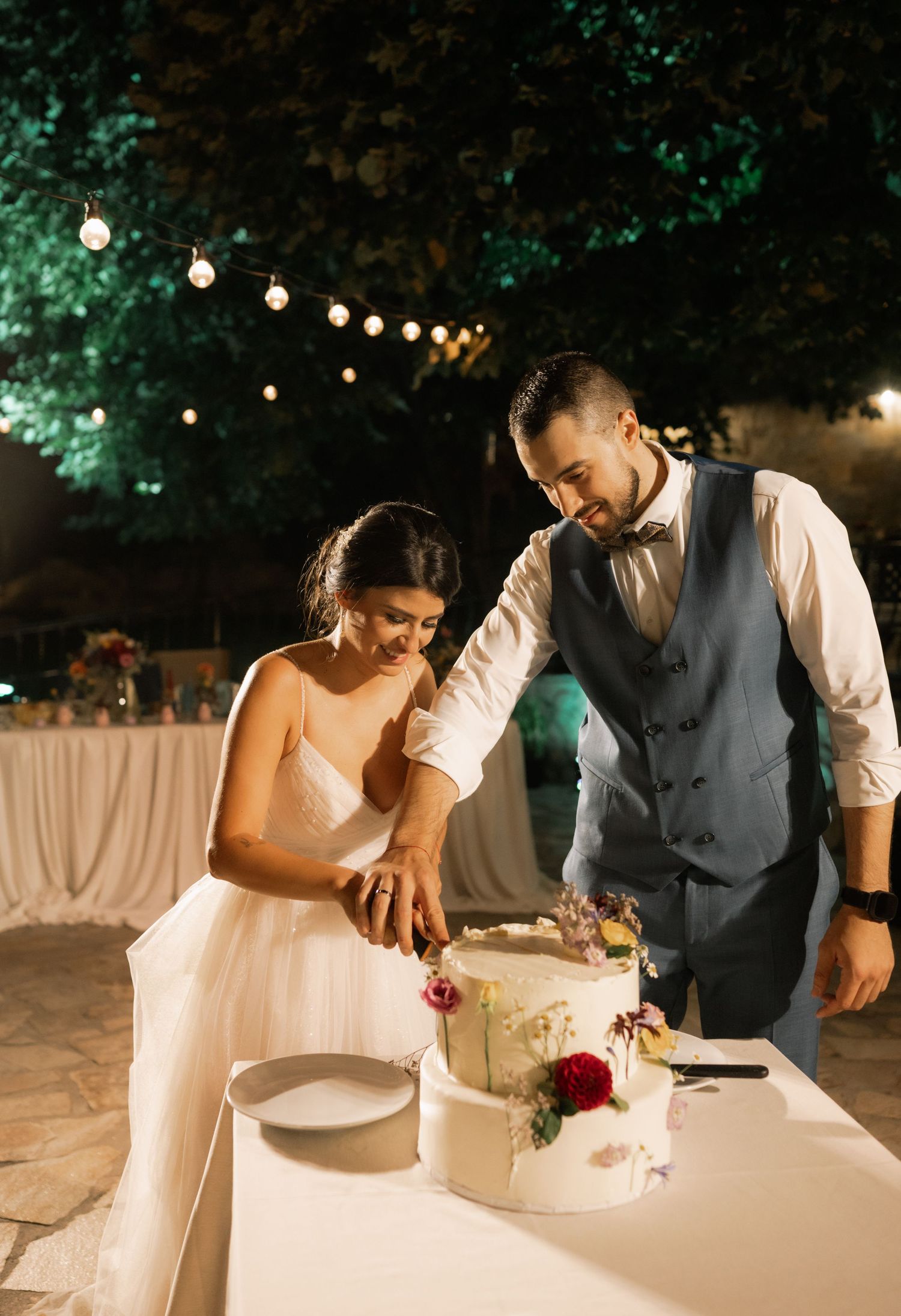 A couple in wedding attire cuts their white wedding cake together during an outdoor evening reception with string lights.