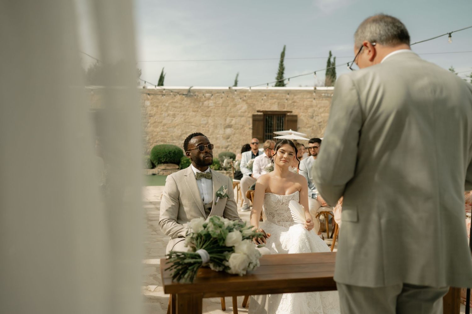 Interracial couple exchanging vows at an outdoor wedding ceremony with lush floral arrangements and stone venue backdrop.