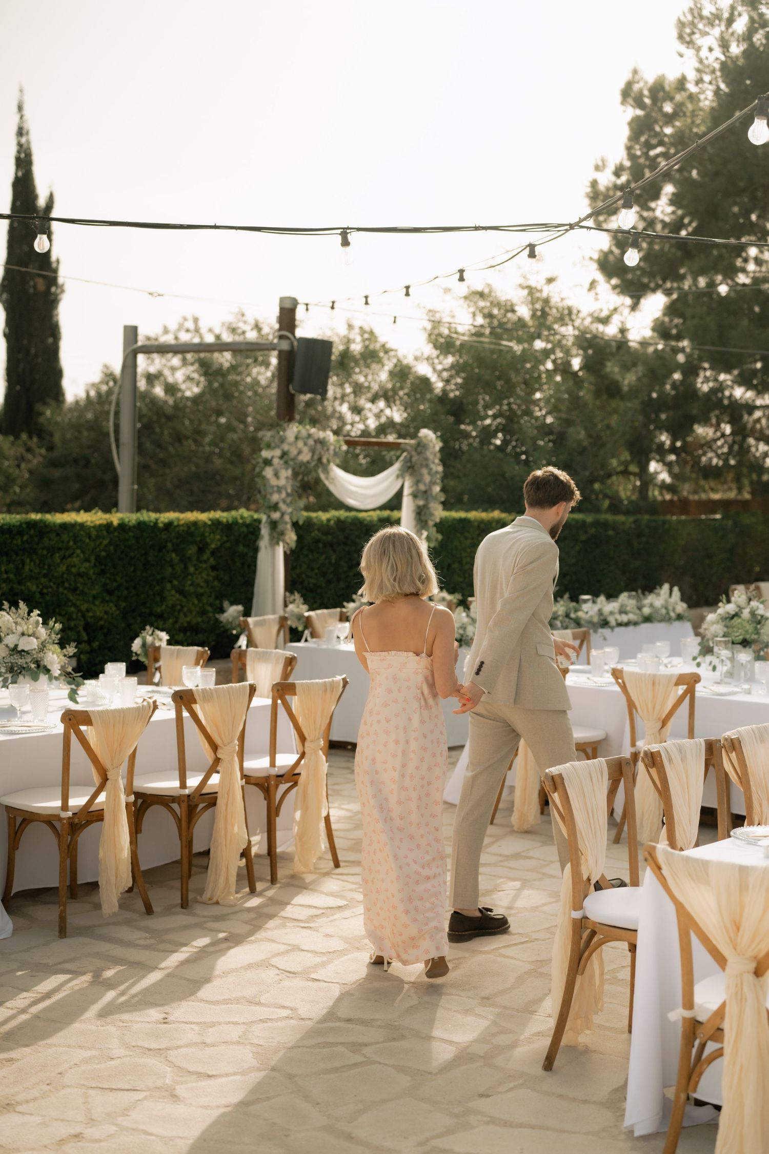 Bride and groom walk through elegant outdoor wedding reception with wooden cross-back chairs and string lights.