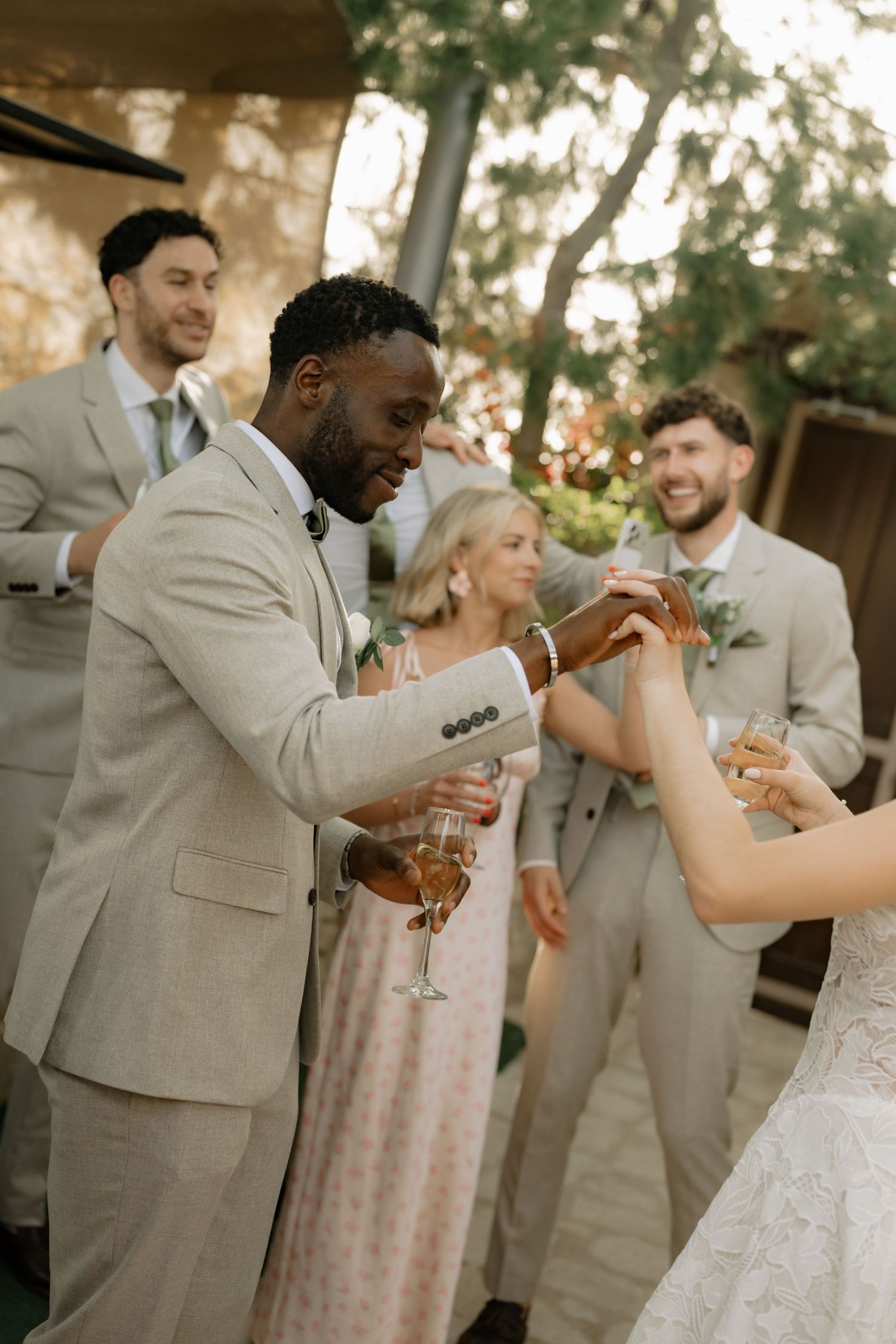 Wedding guests in beige suits toast with champagne glasses outdoors at an elegant garden celebration.